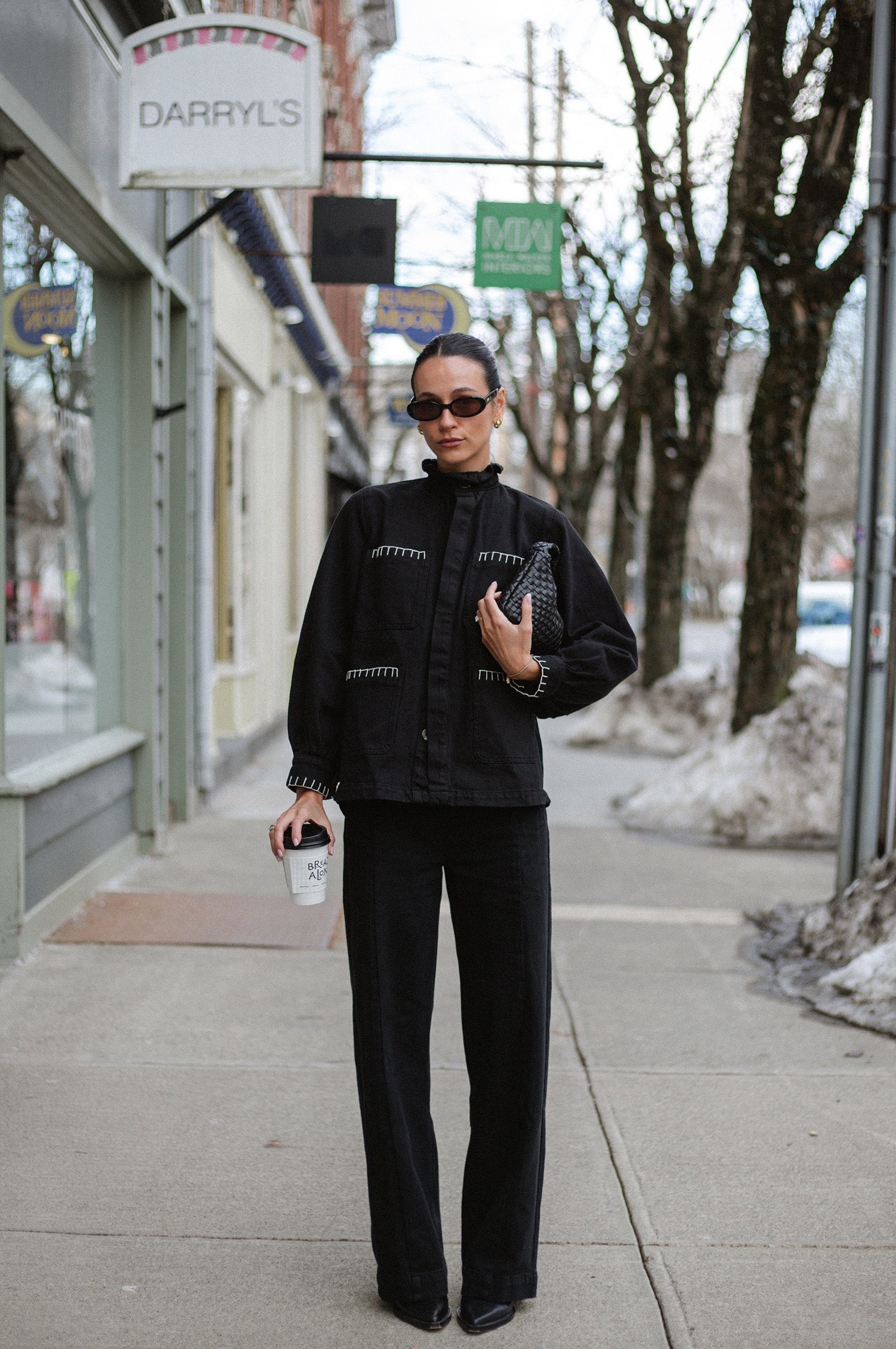 woman holding coffee wearing a denim jacket with white stitching and black high rise, wide leg jeans with a sailor button front fastening detail outside