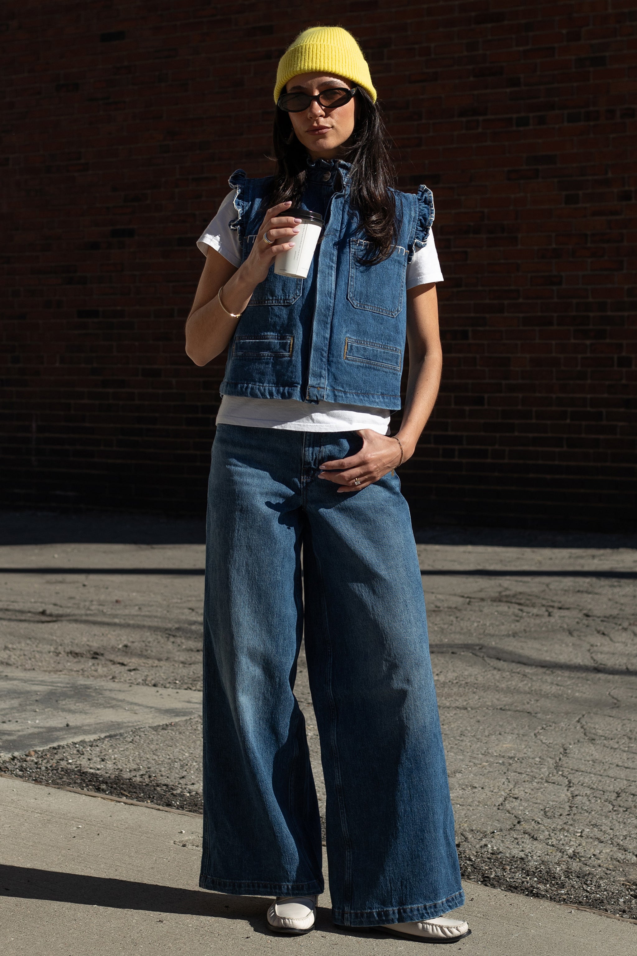 Woman holding a coffee cup and wearing a denim vest with frill details, white shirt, and blue wide leg jeans against a brick wall.