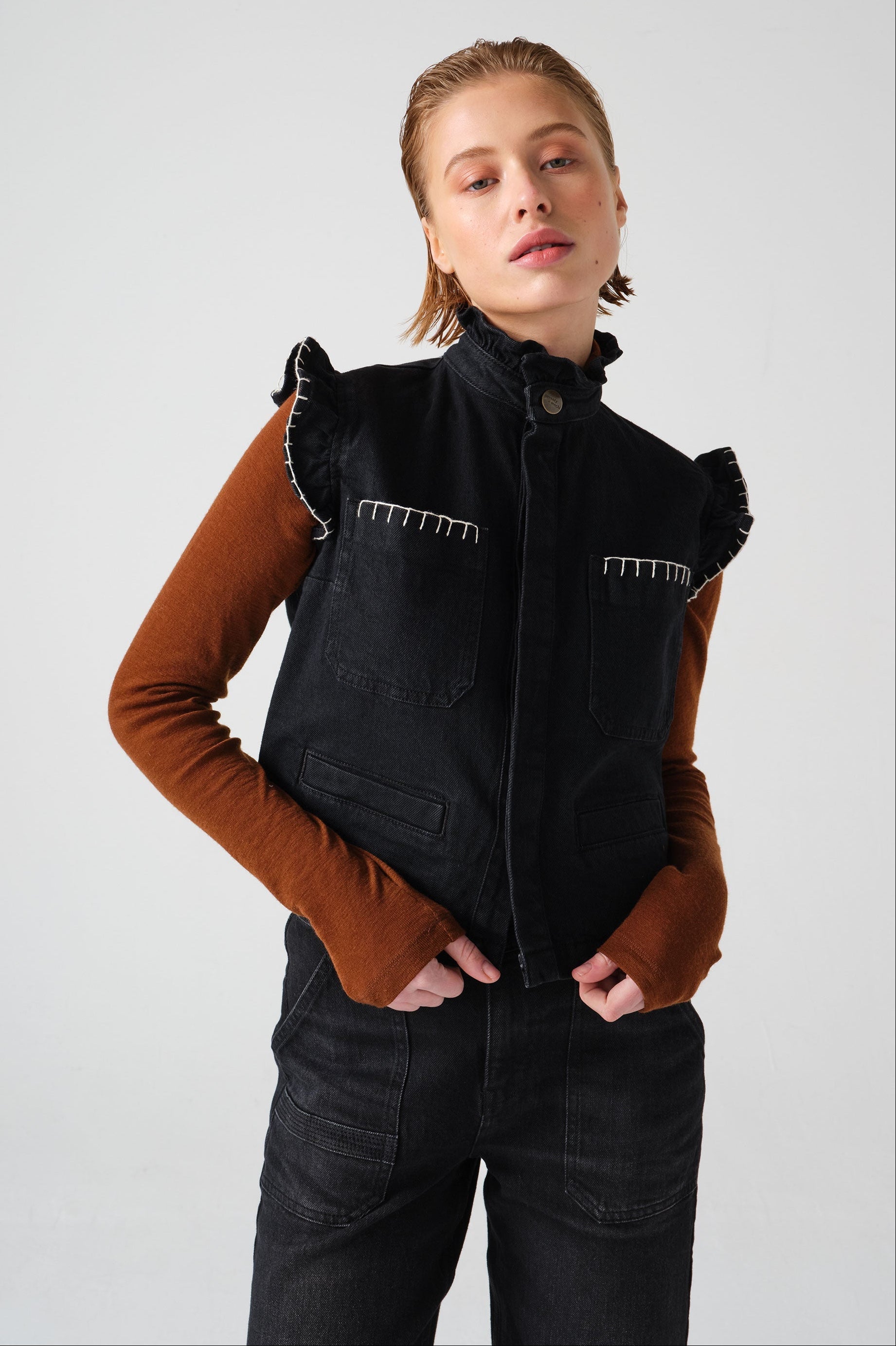 woman wearing a black vest/ waistcoat with a frill collar and our signature white stitching on two large patch pockets, paired with a brown top and black jeans against a white backdrop
