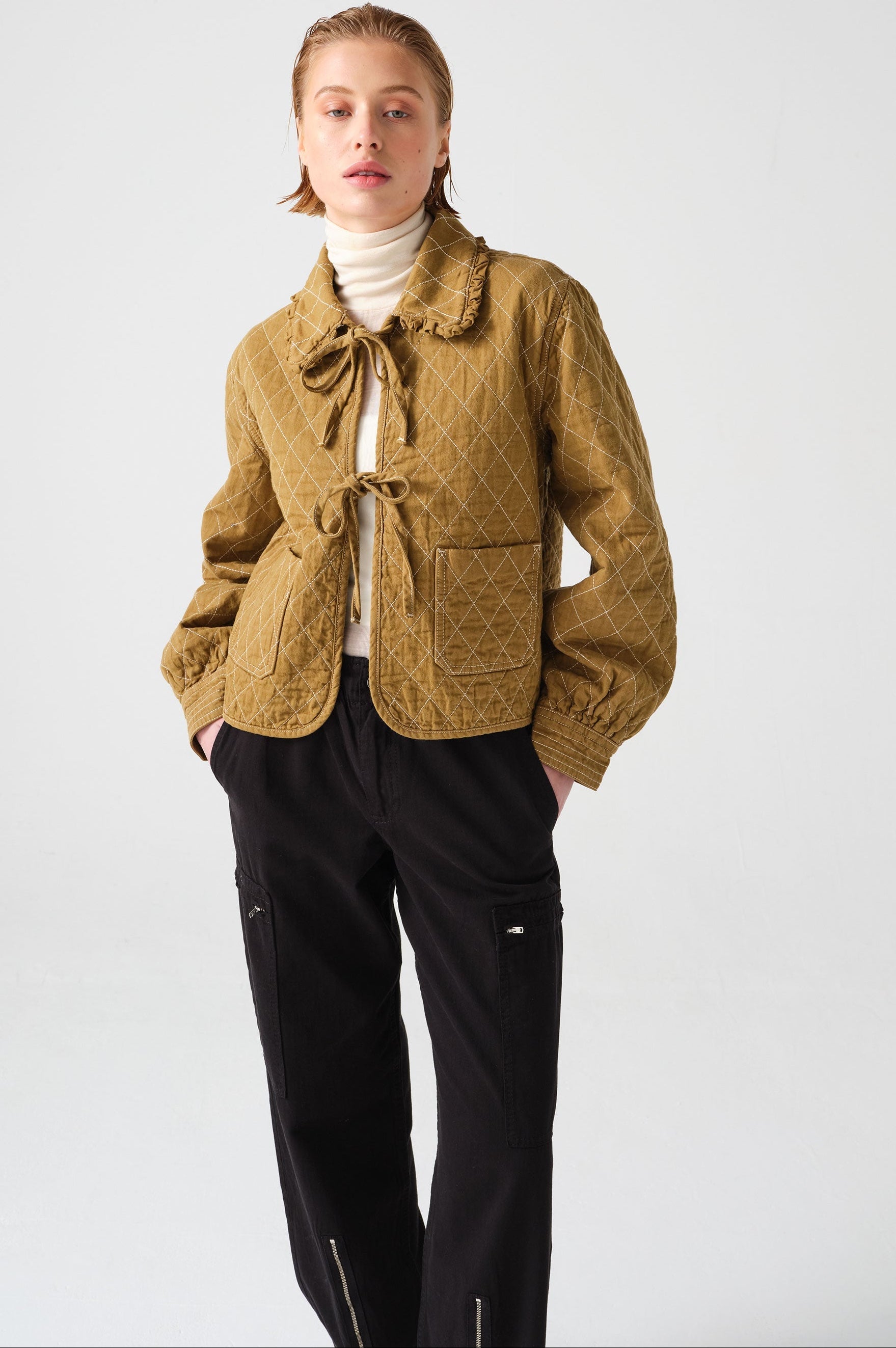 woman wearing a pair of black, lightweight, cargo style pants with an elastic waist, deep zip pockets, and a zip detailed hem paired with a white top, olive quilted jacket, and black boots against a white backdrop