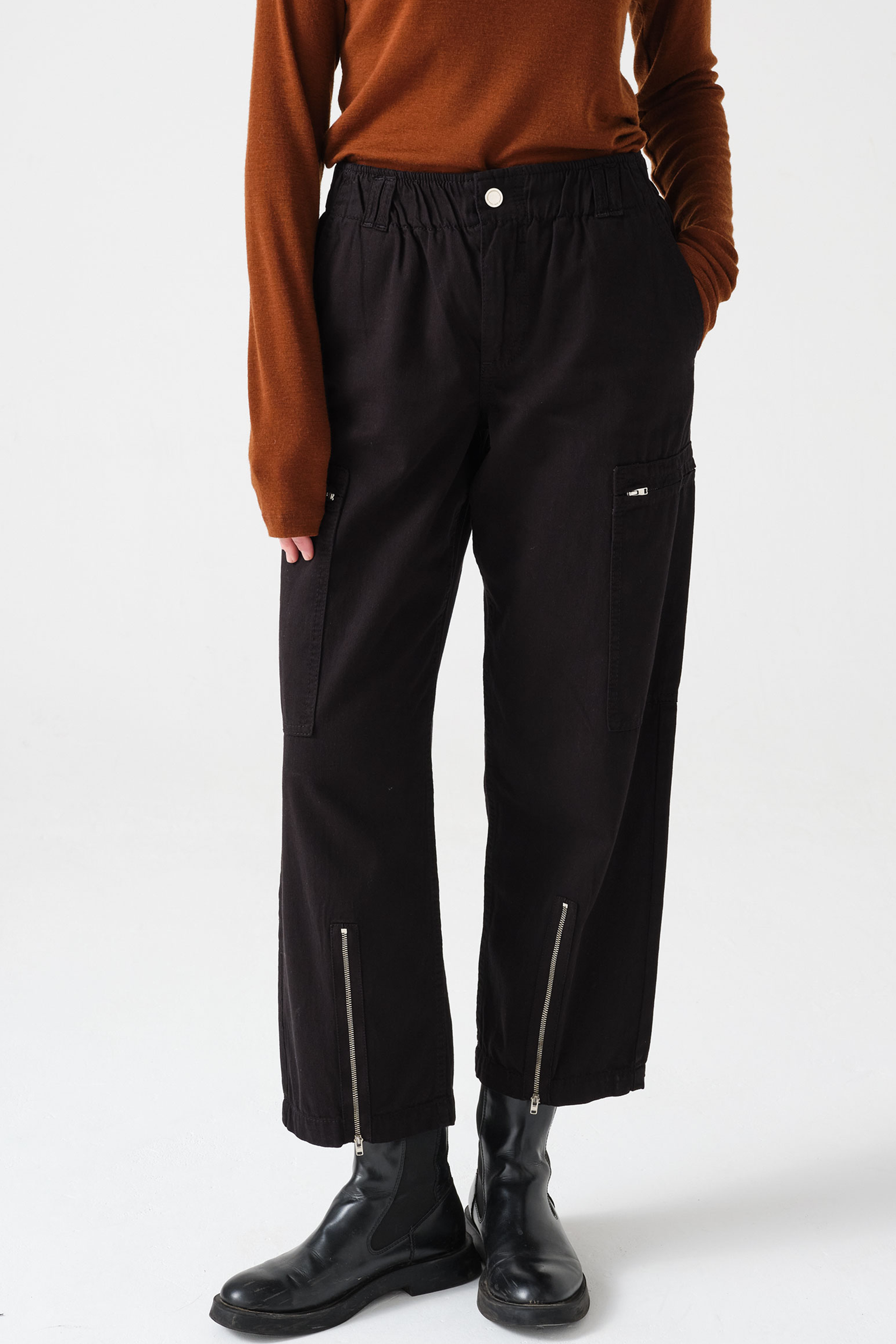woman wearing a pair of black, lightweight, cargo style pants with an elastic waist, deep zip pockets, and a zip detailed hem paired with an orange top and black boots against a white backdrop