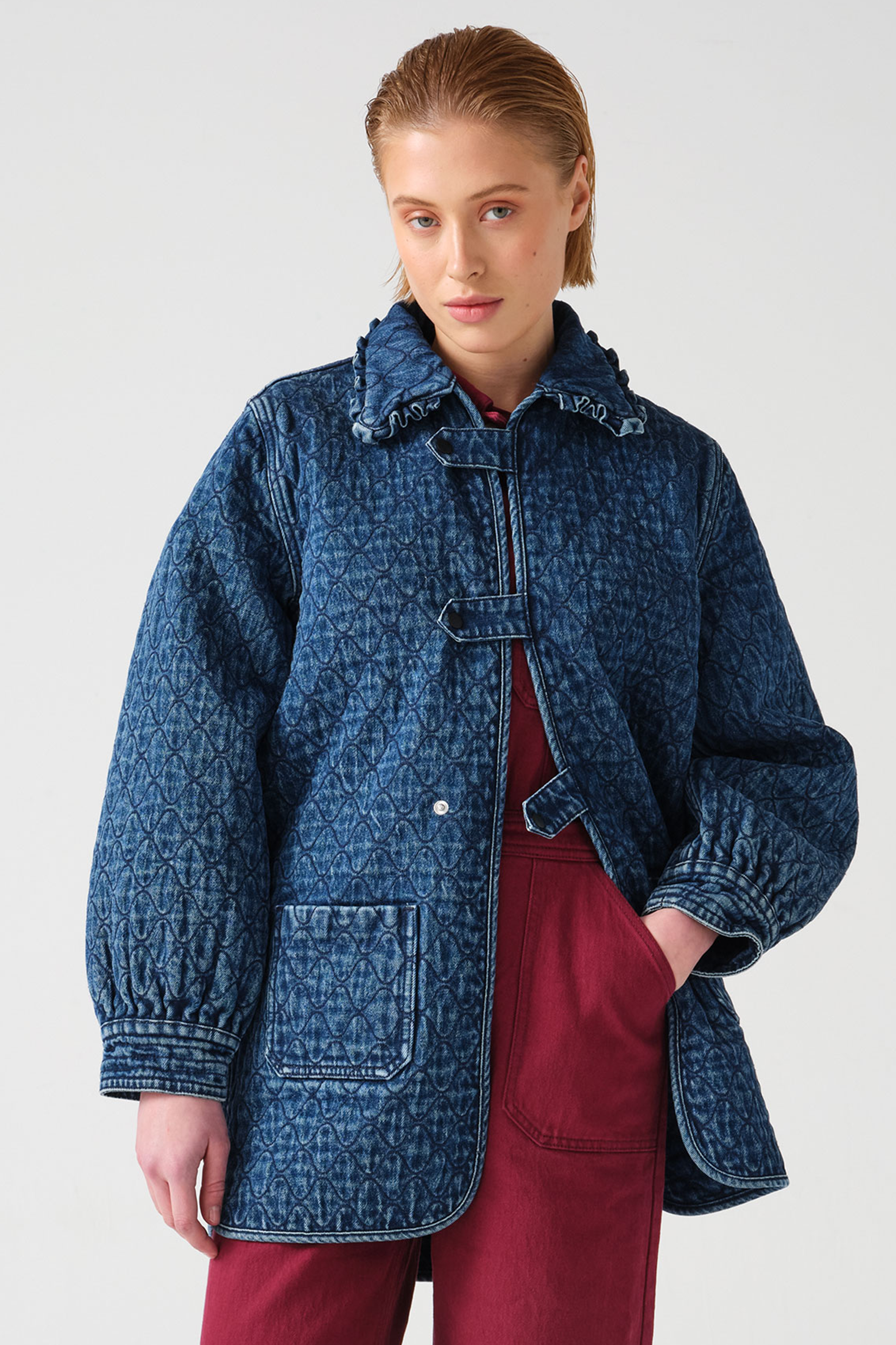 woman wearing a blue quilted jacket with a frill collar, tab closures, and two large pockets with red overalls on a white background