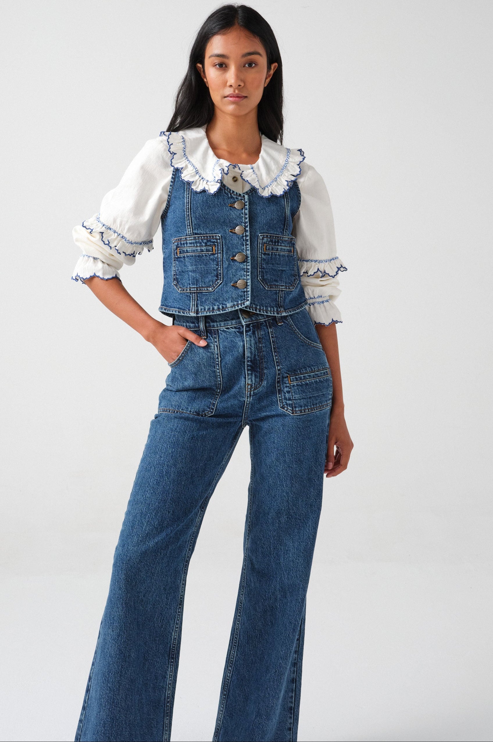 woman wearing an organic cotton blouse with electric blue frill embroidery on the collar and sleeves, paired with blue jeans and a blue denim vest/ waistcoat