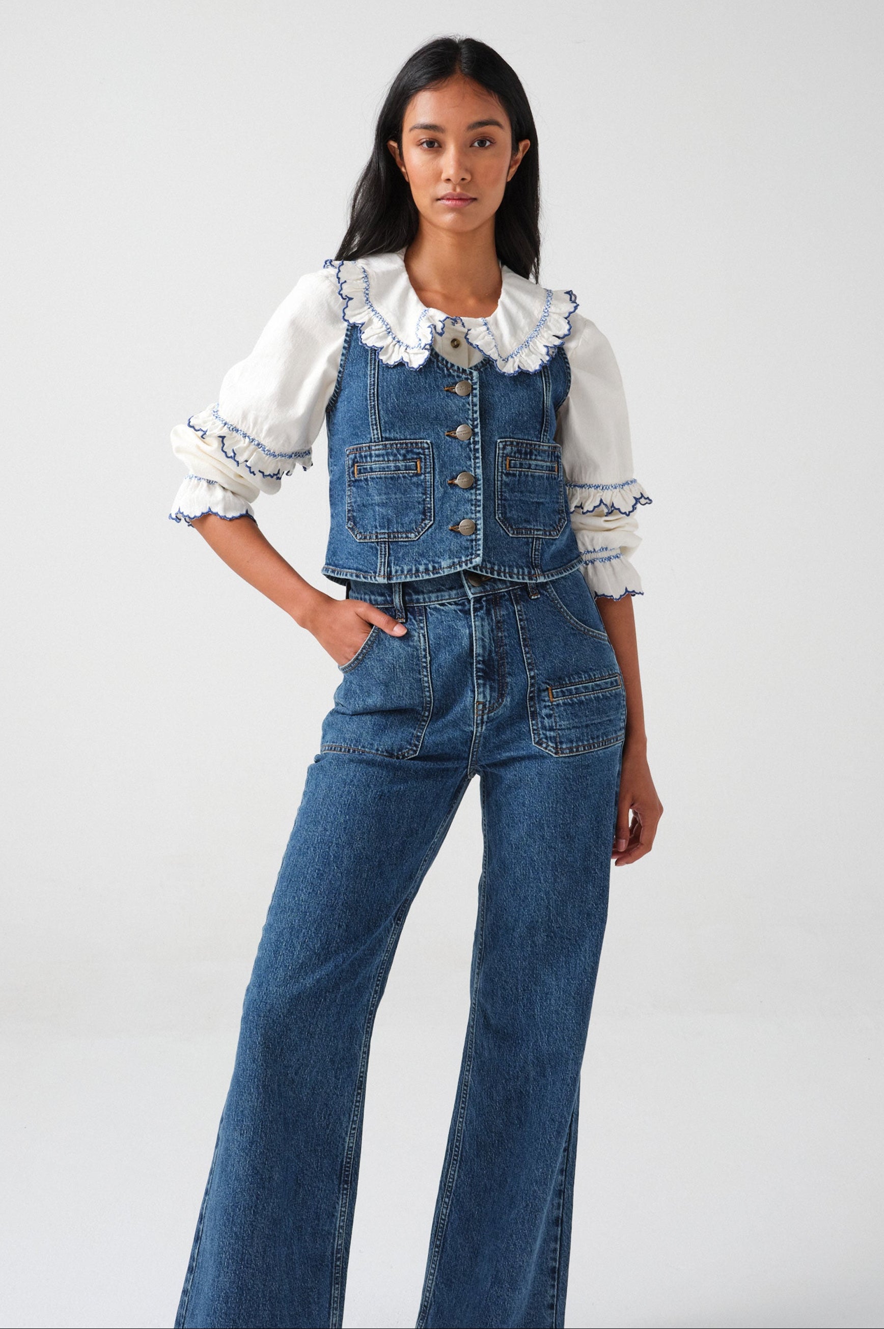 woman wearing an organic cotton blouse with electric blue frill embroidery on the collar and sleeves, paired with blue jeans and a blue denim vest/ waistcoat