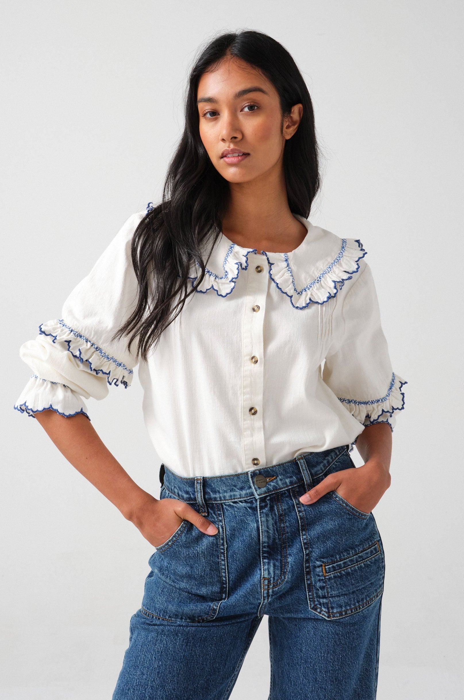 woman wearing an organic cotton blouse with electric blue frill embroidery on the collar and sleeves, paired with blue jeans.