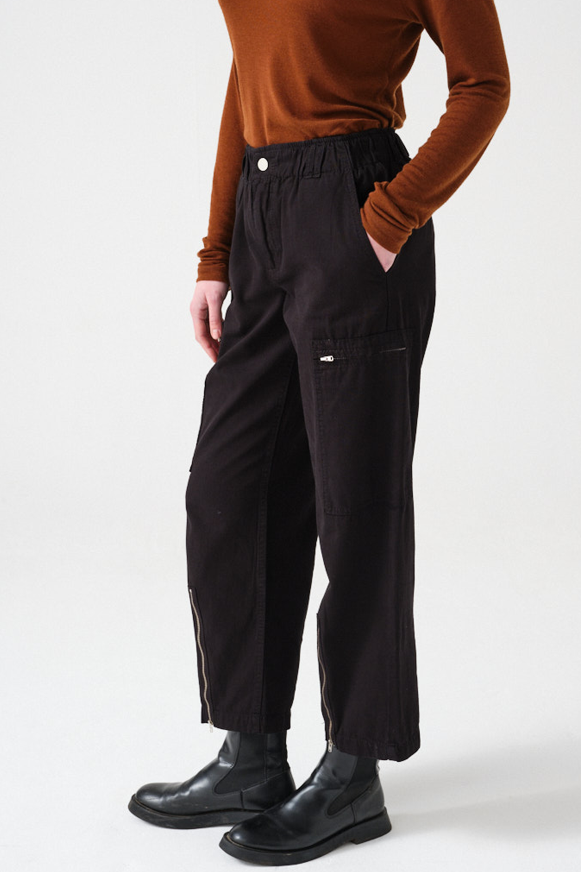 woman wearing a pair of black, lightweight, cargo style pants with an elastic waist, deep zip pockets, and a zip detailed hem paired with an orange top and black boots against a white backdrop
