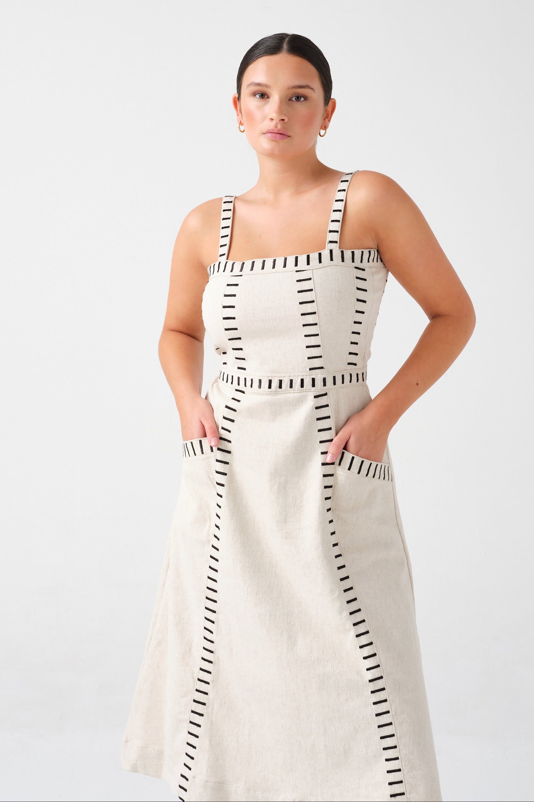 A woman wearing an ecru midi denim dress with black stitch details, a square neckline, front pockets, and an open, lace-up back on a white background