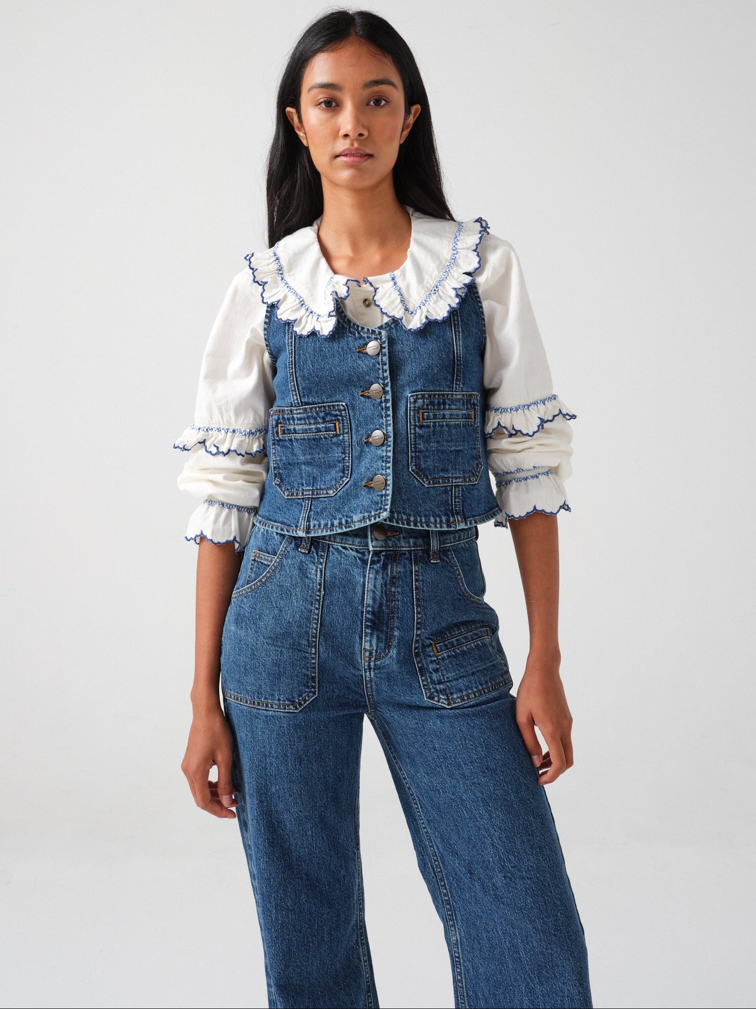 woman wearing an organic cotton blouse with electric blue frill embroidery on the collar and sleeves, paired with blue jeans and a blue denim vest/ waistcoat