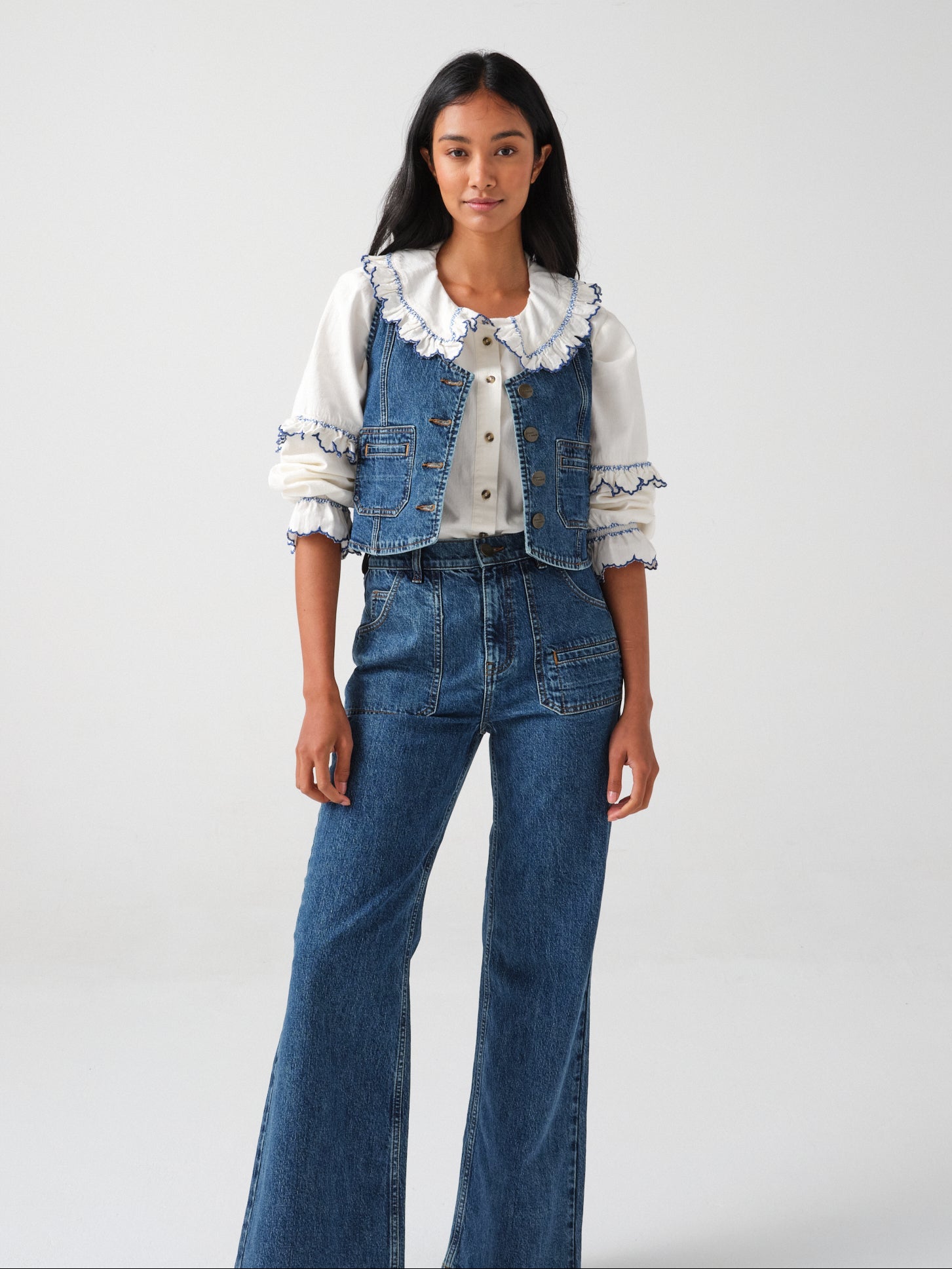 woman wearing an organic cotton blouse with electric blue frill embroidery on the collar and sleeves, paired with blue jeans and a blue denim vest/ waistcoat