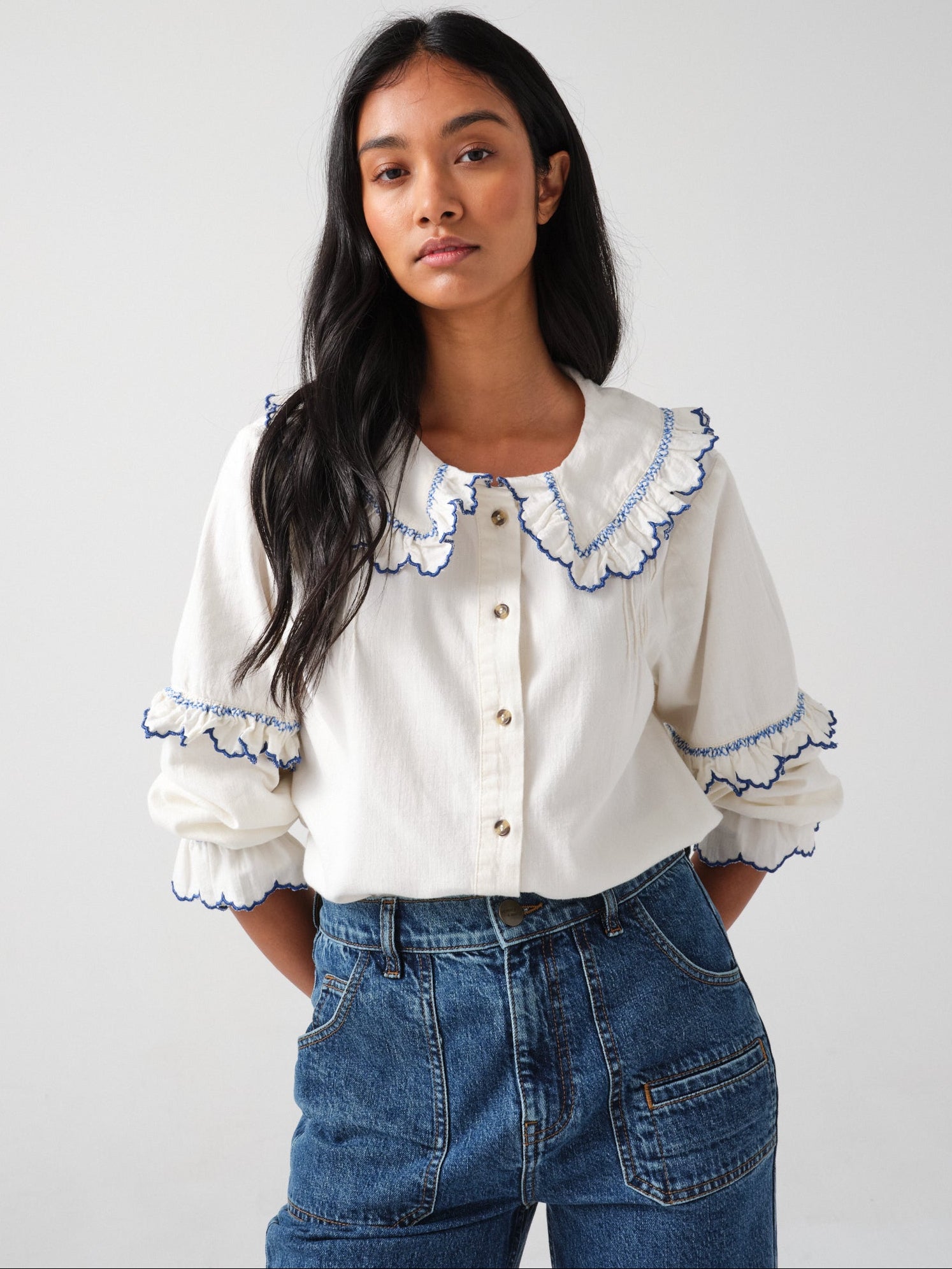 woman wearing an organic cotton blouse with electric blue frill embroidery on the collar and sleeves, paired with blue jeans on a white background