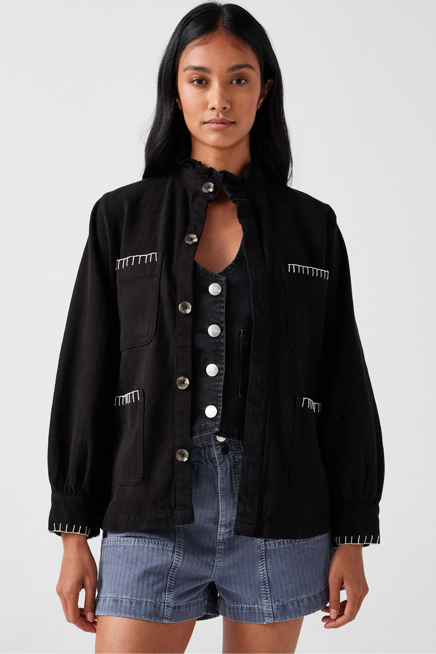 A woman wearing a black denim jacket with a frill collar and our signature white blanket stitching on four large patch pockets, paired with blue shorts and a black vest against a white backdrop