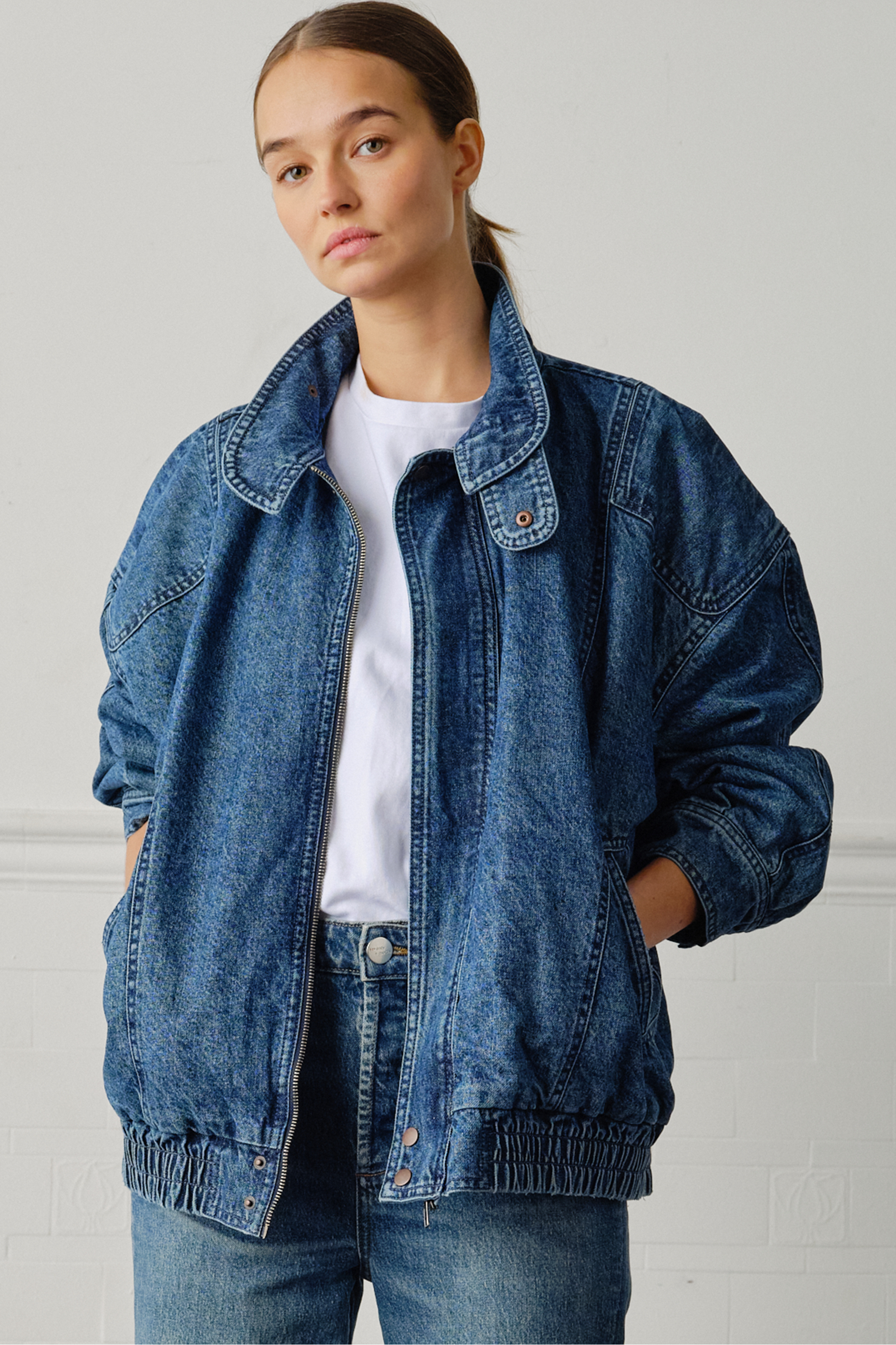 A woman wearing an oversized padded and seamed blue denim retro style bomber jacket with a round neckline, zip, and button closures at the top, bottom, and collar with a white top and blue jeans against a white backdrop