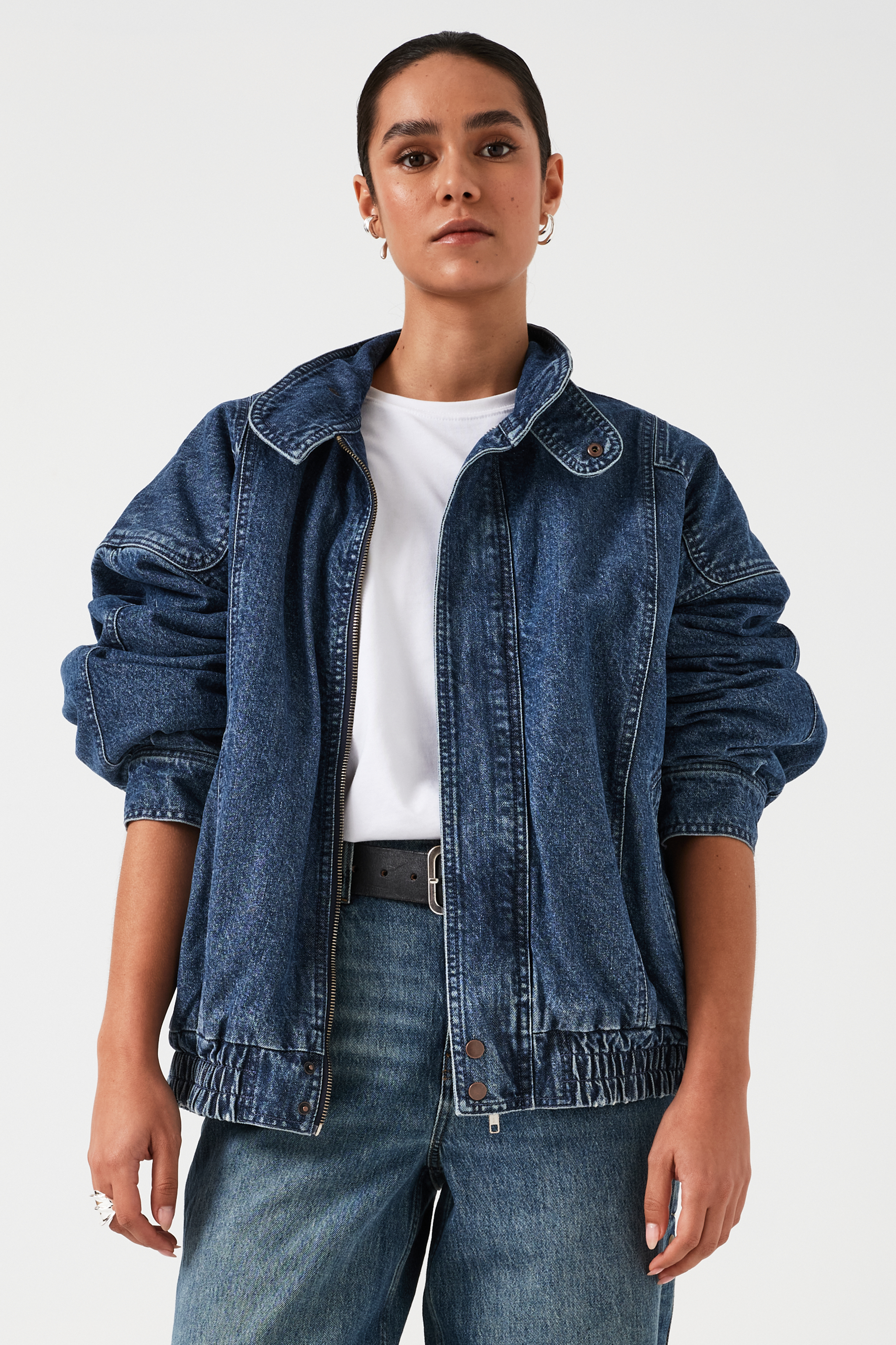 A woman wearing an oversized padded and seamed blue denim retro style bomber jacket with a round neckline, zip, and button closures at the top, bottom, and collar with a white top and blue jeans against a white backdrop
