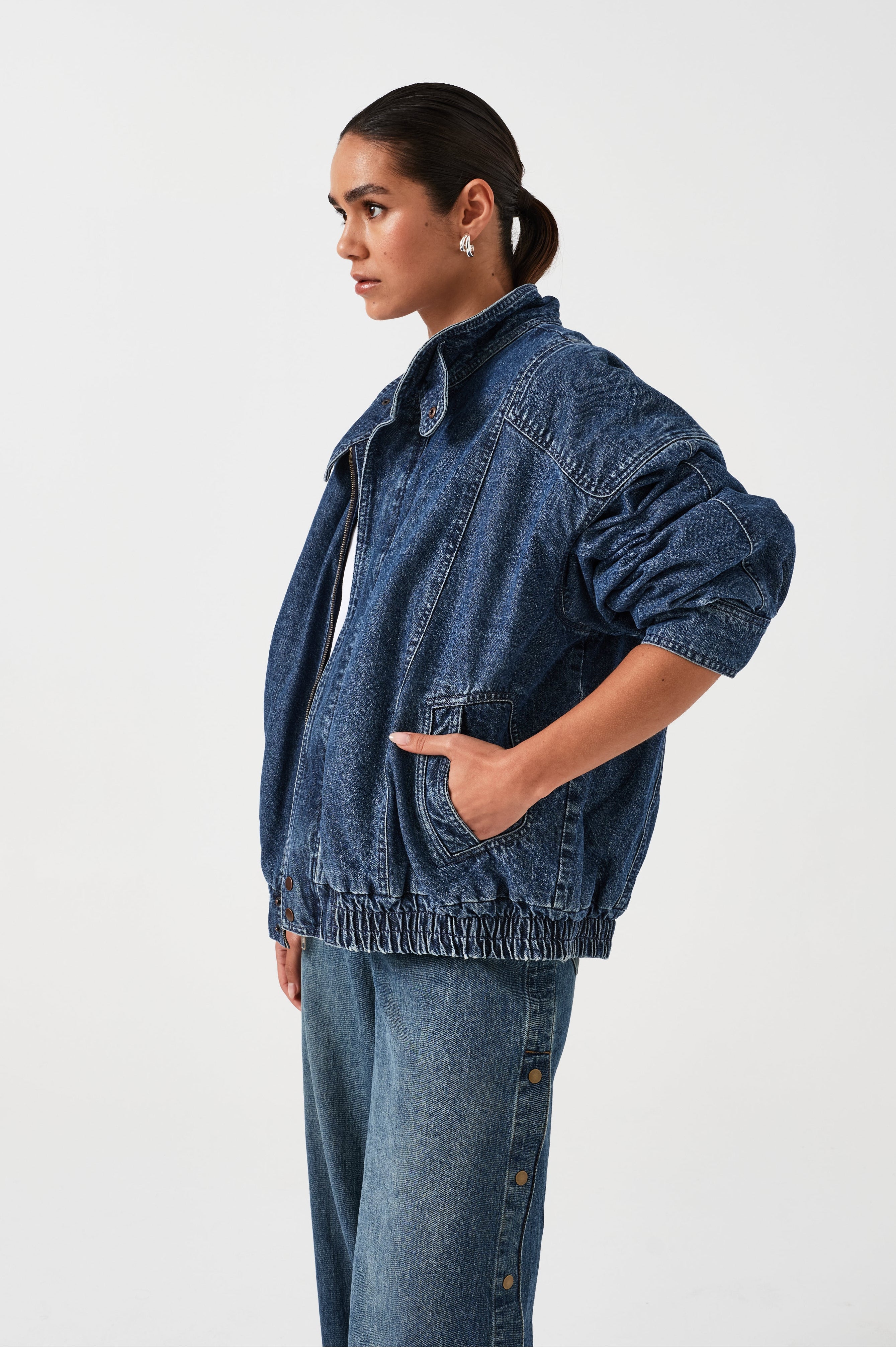 A woman wearing an oversized padded and seamed blue denim retro style bomber jacket with a round neckline, zip, and button closures at the top, bottom, and collar with a white top and blue jeans against a white backdrop