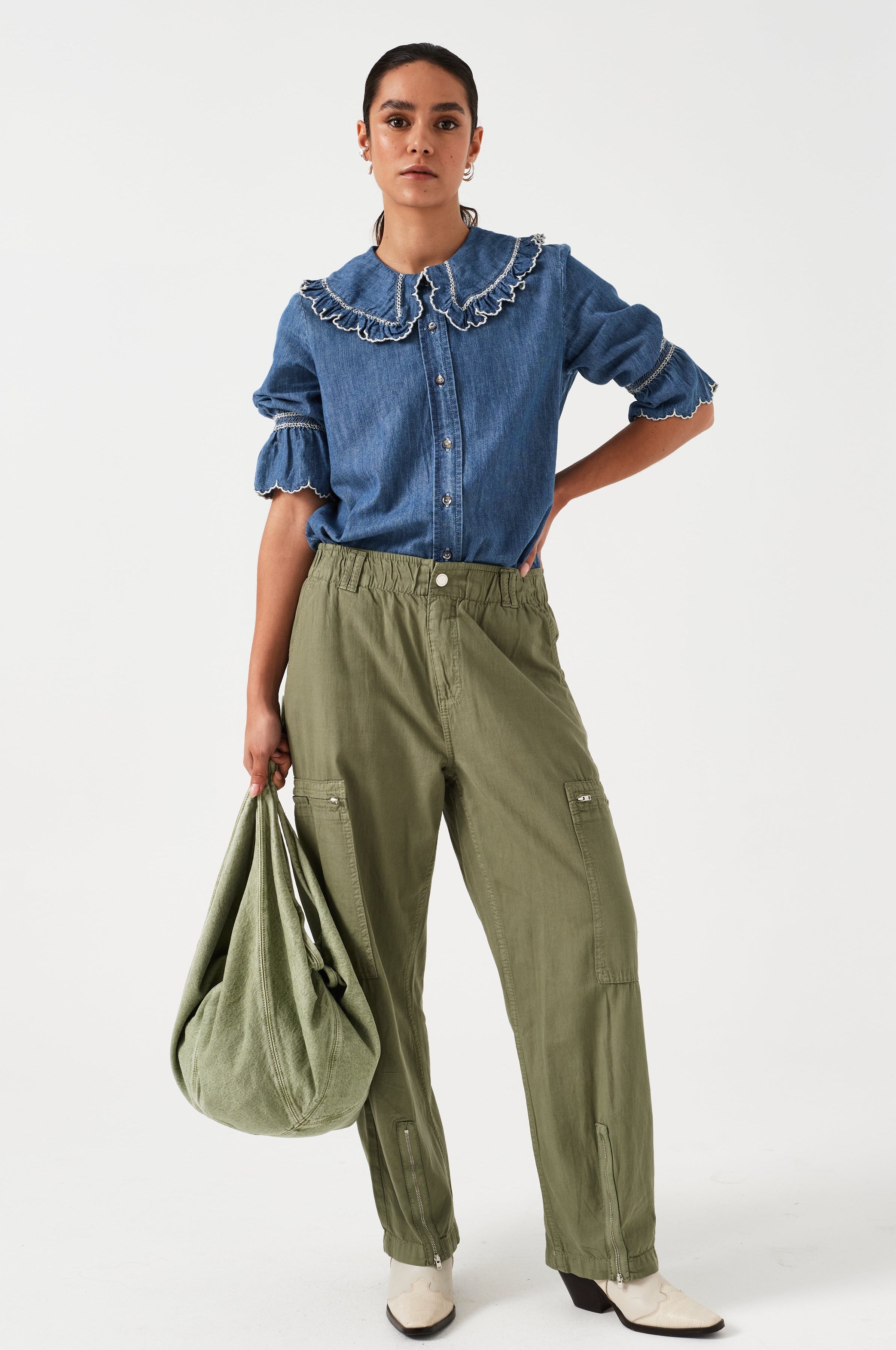 woman wearing an indigo denim blouse with white embroidery frill on the collar and sleeves and khaki pants with a khaki bag on a white background