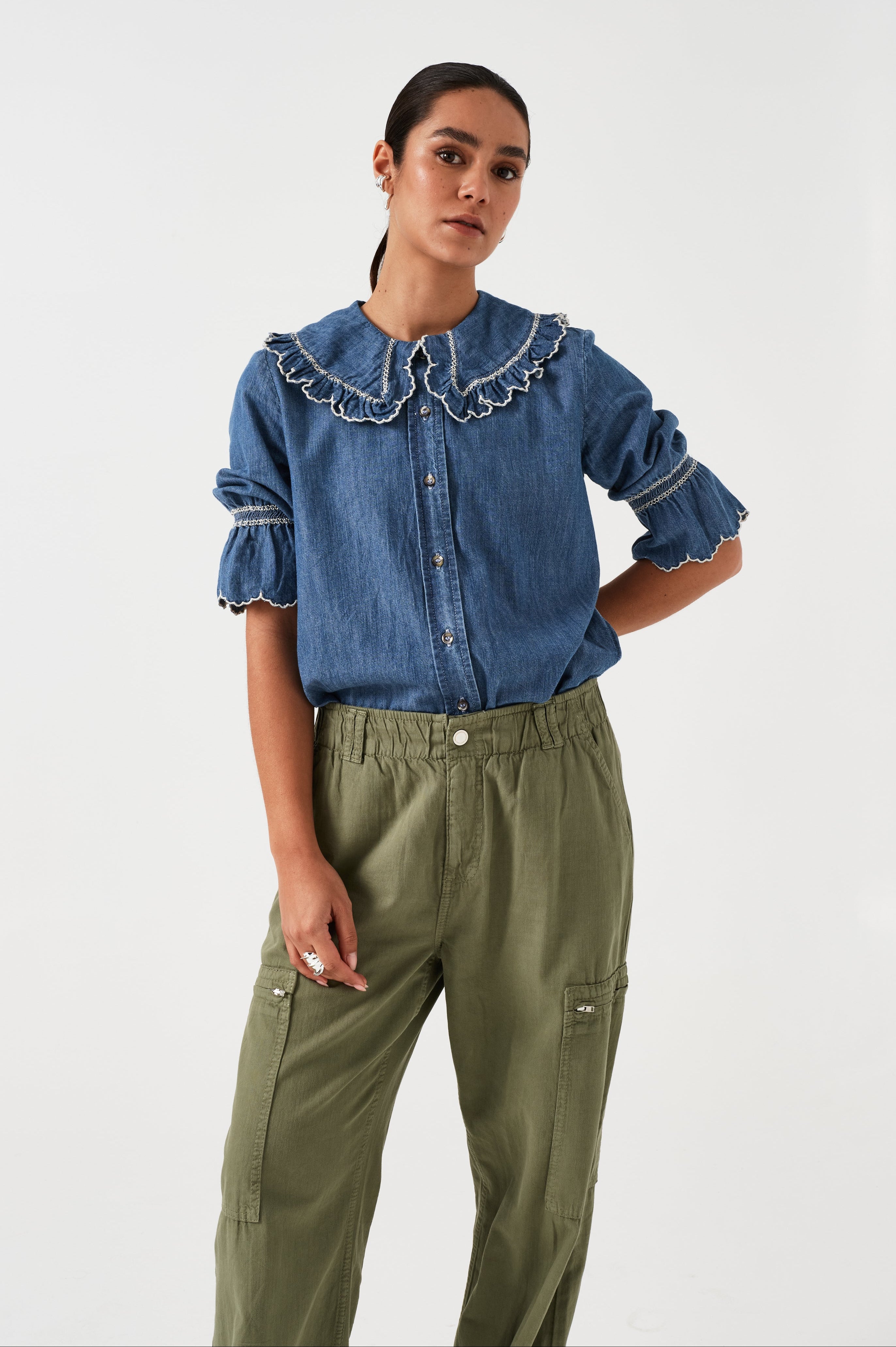 woman wearing an indigo denim blouse with white embroidery frill on the collar and sleeves and khaki pants on a white background