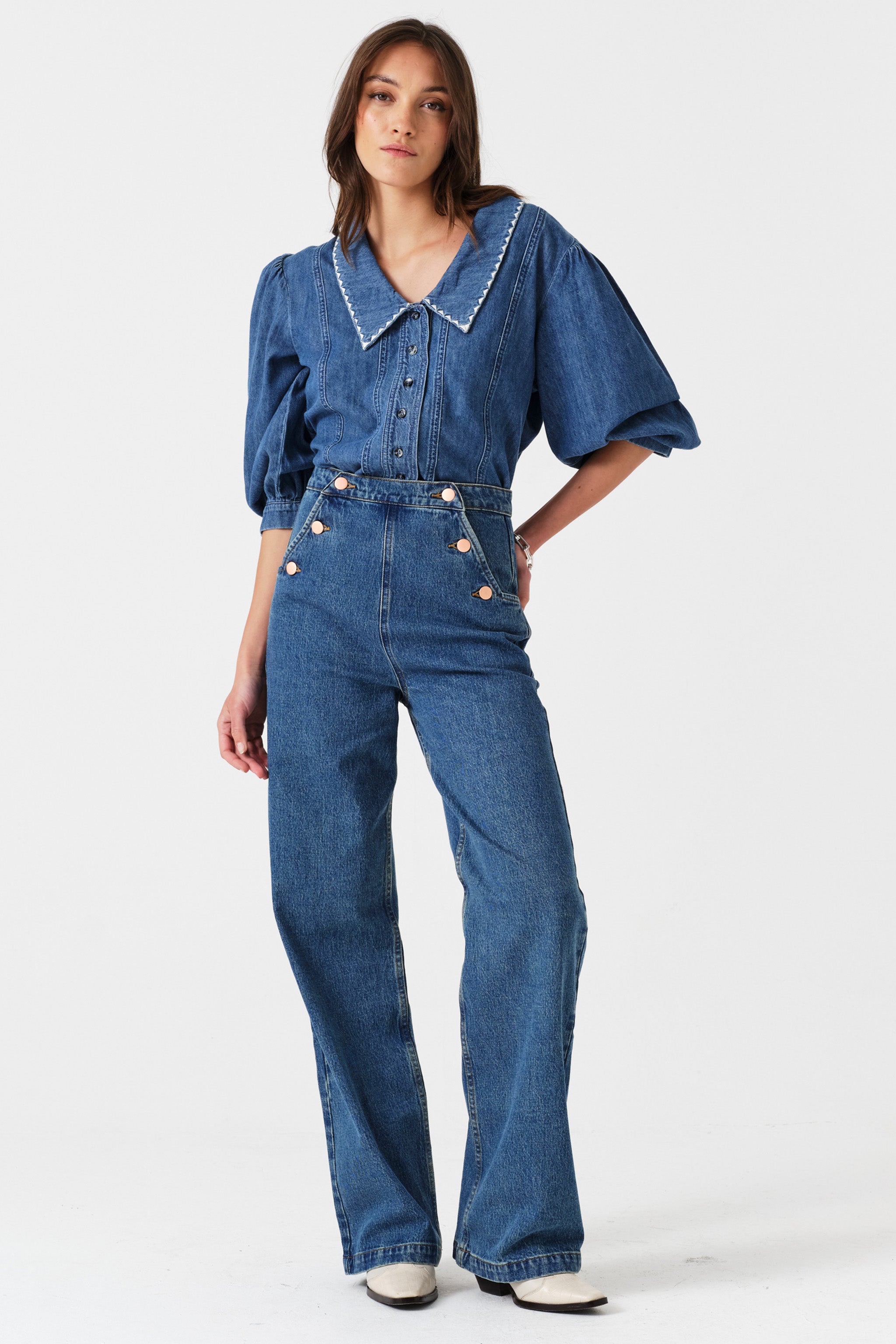 Woman wearing a blue denim button up top with high waisted jeans on a white background.