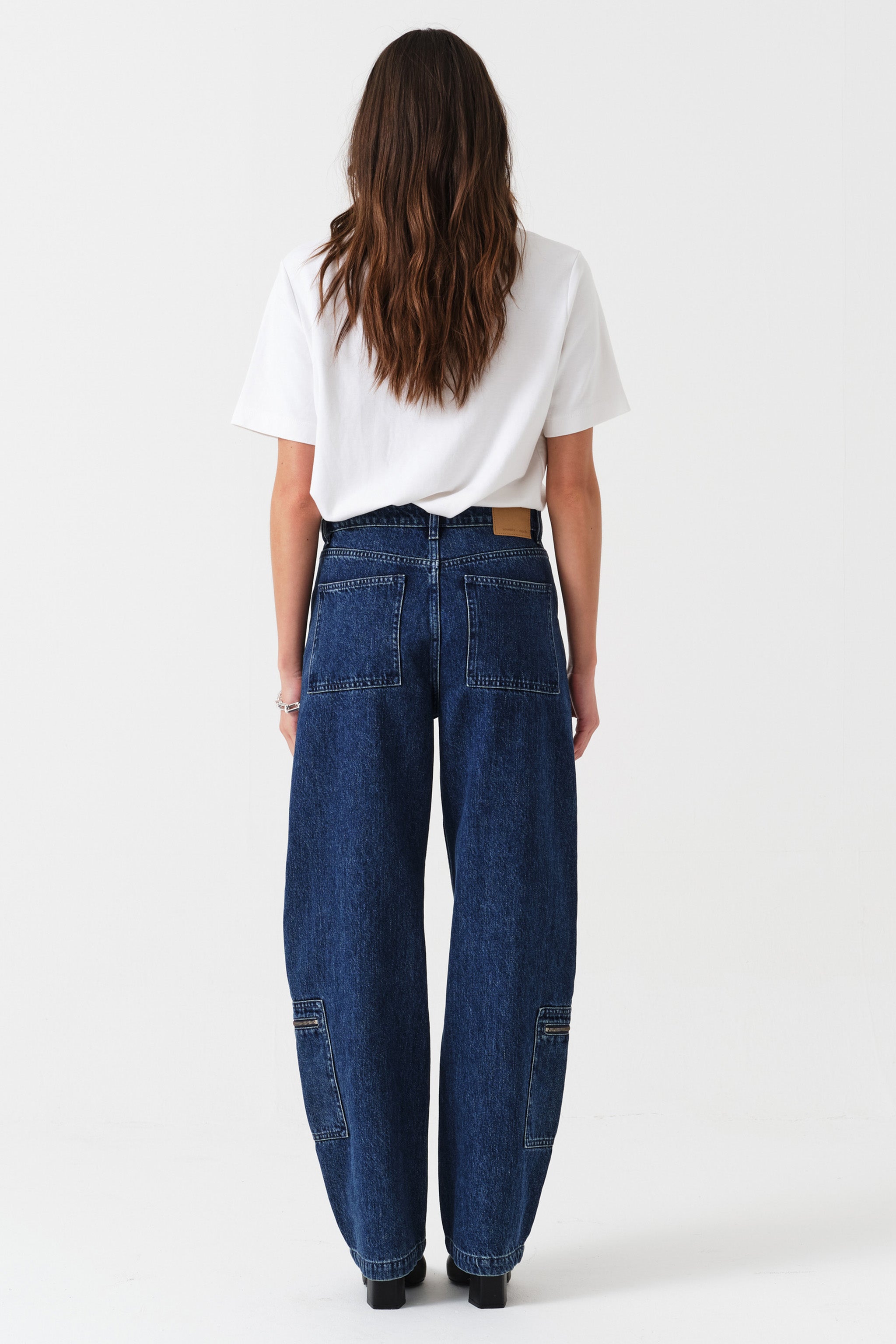 Woman wearing a white t-shirt and blue jeans with zip details on a white background photographed from the
back