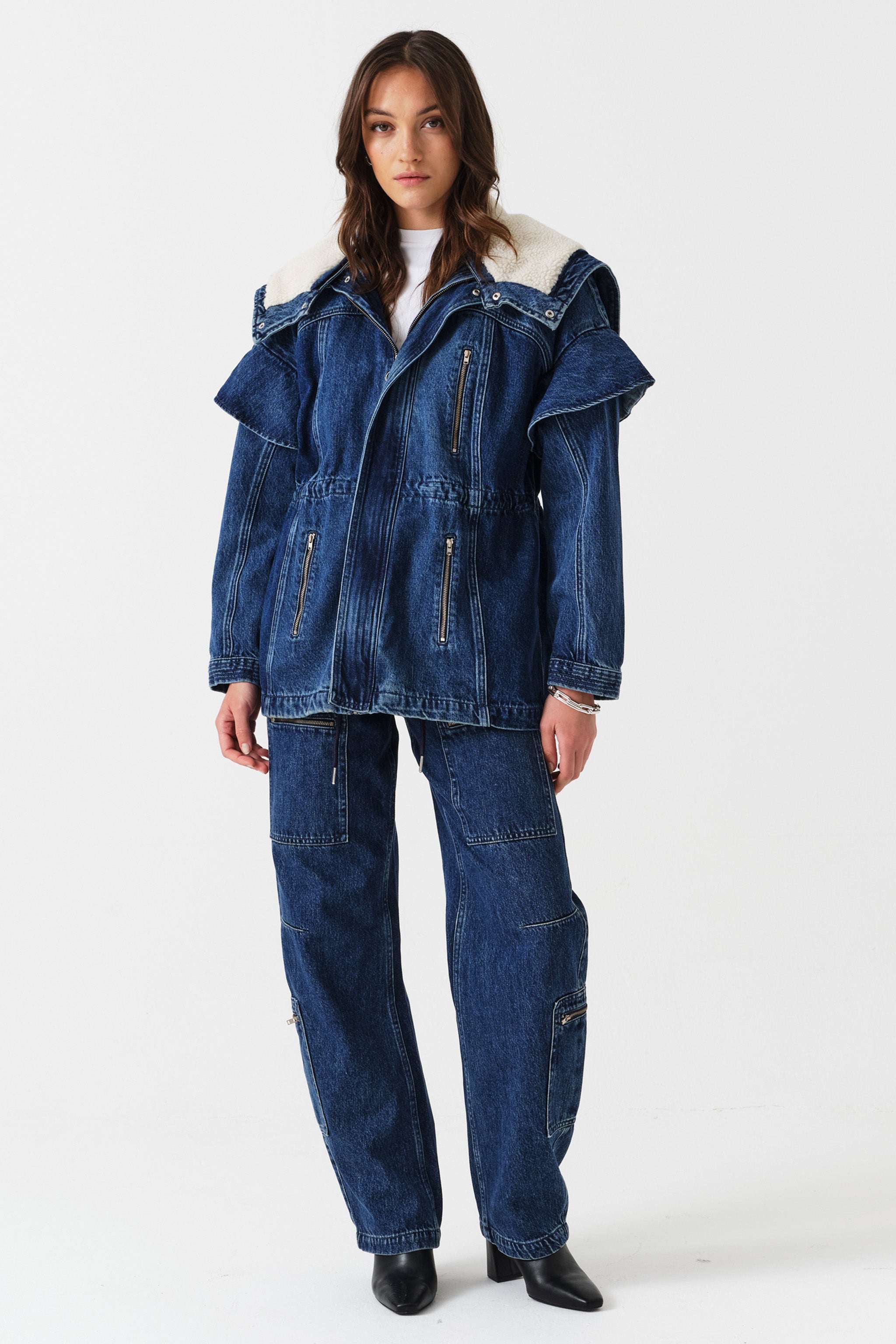 Woman wearing a white t-shirt, blue denim jacket with sherpa collar and zip details, and blue jeans with zip details on a white background