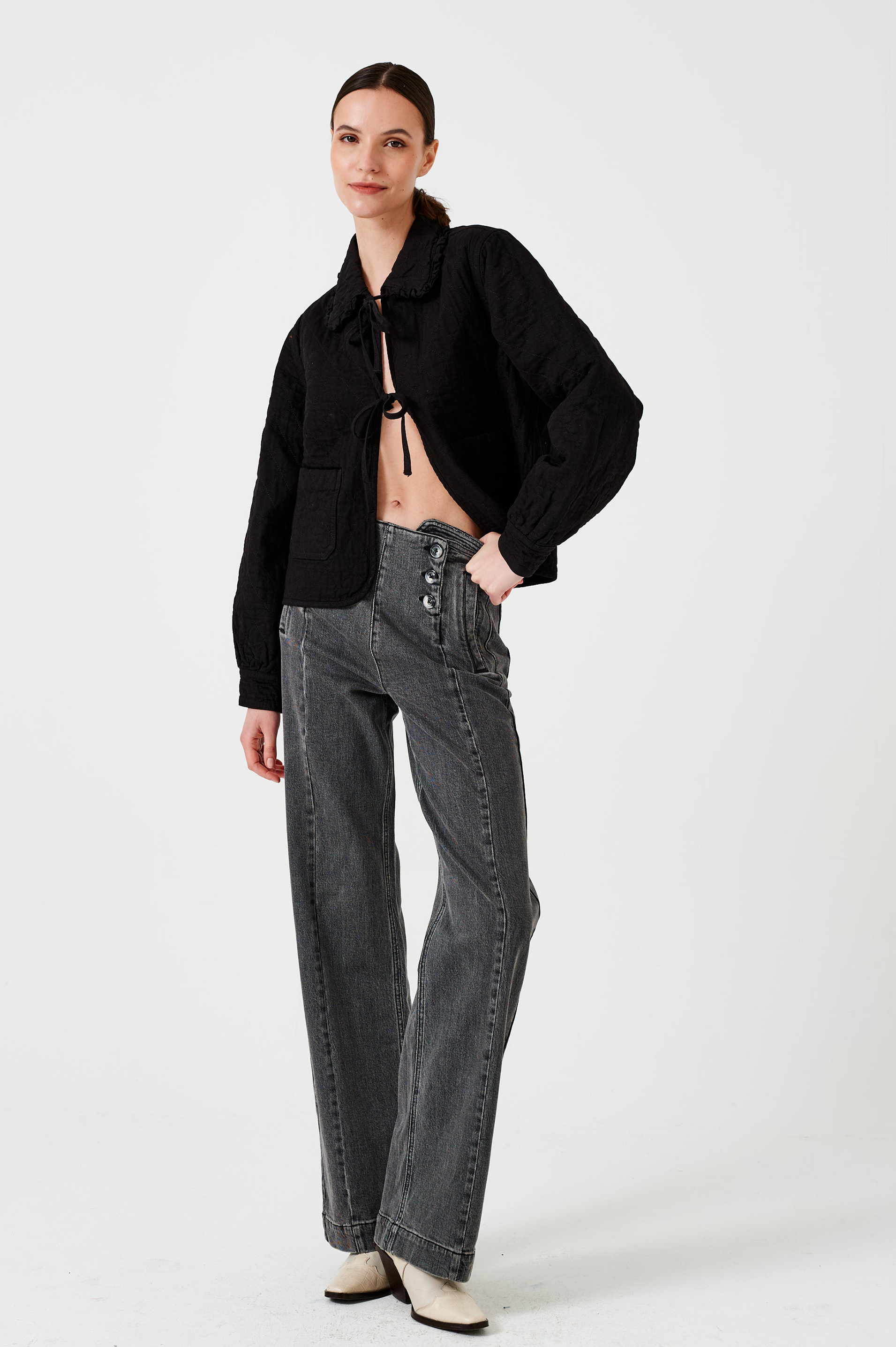Woman wearing a black quilted denim jacket with a ruffled collar, front tie closures, and two large pockets, and black jeans on a white background