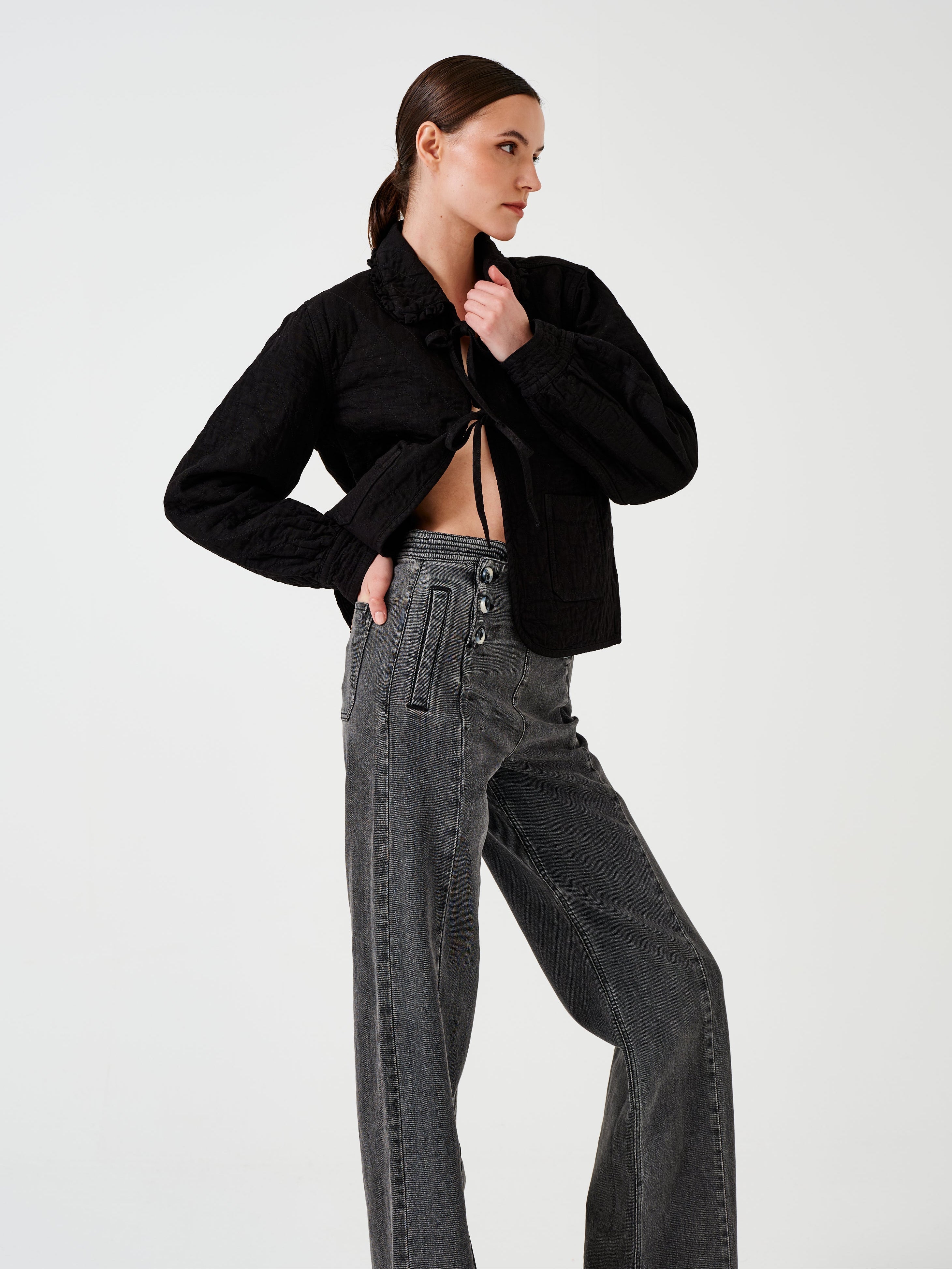 Woman wearing a black quilted denim jacket with a ruffled collar, front tie closures, and two large pockets, and black jeans on a white background