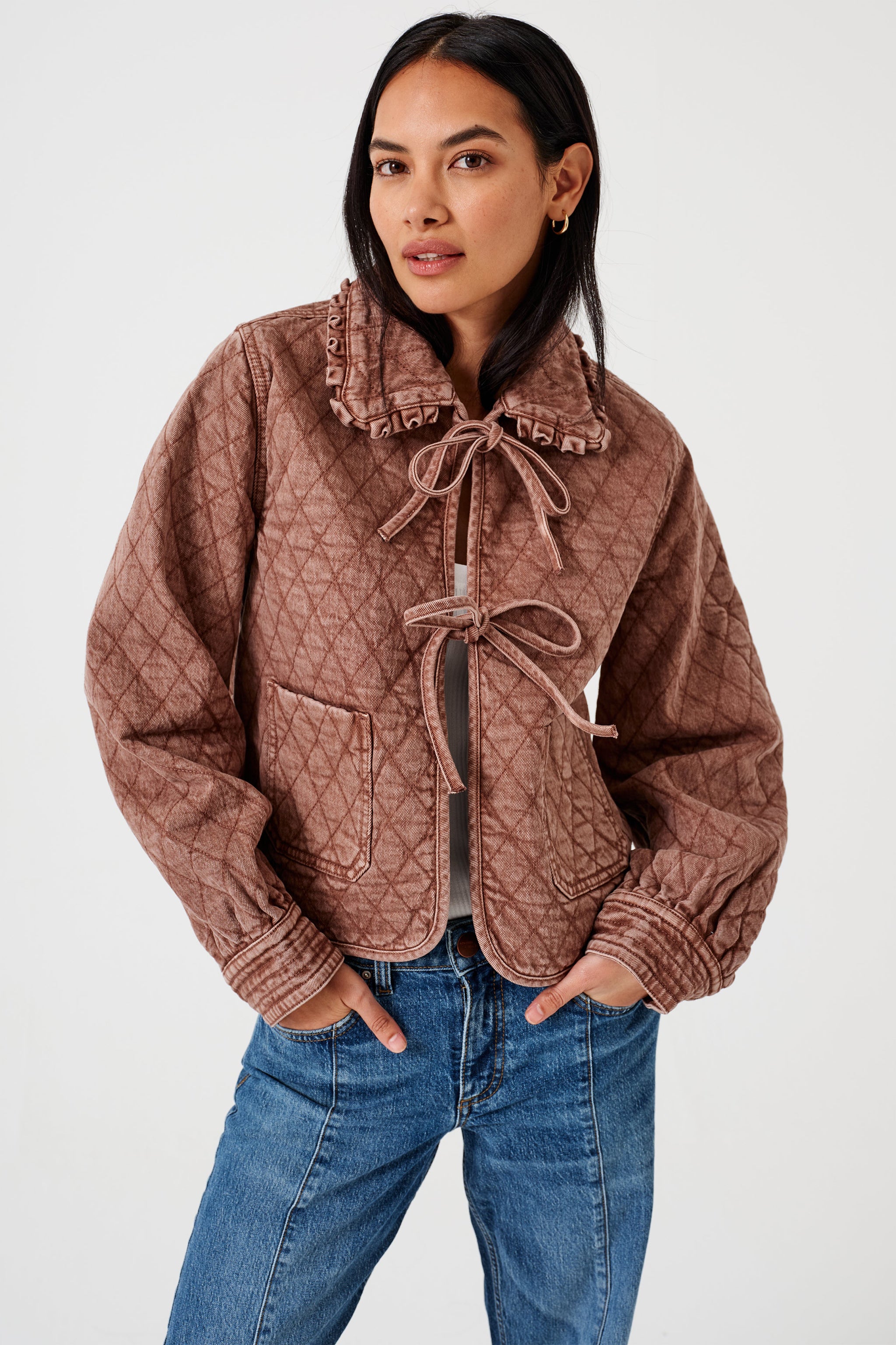 Woman wearing a brown quilted denim jacket with large pockets, a frill collar, and front tie closure, paired with a blue jeans on a white background