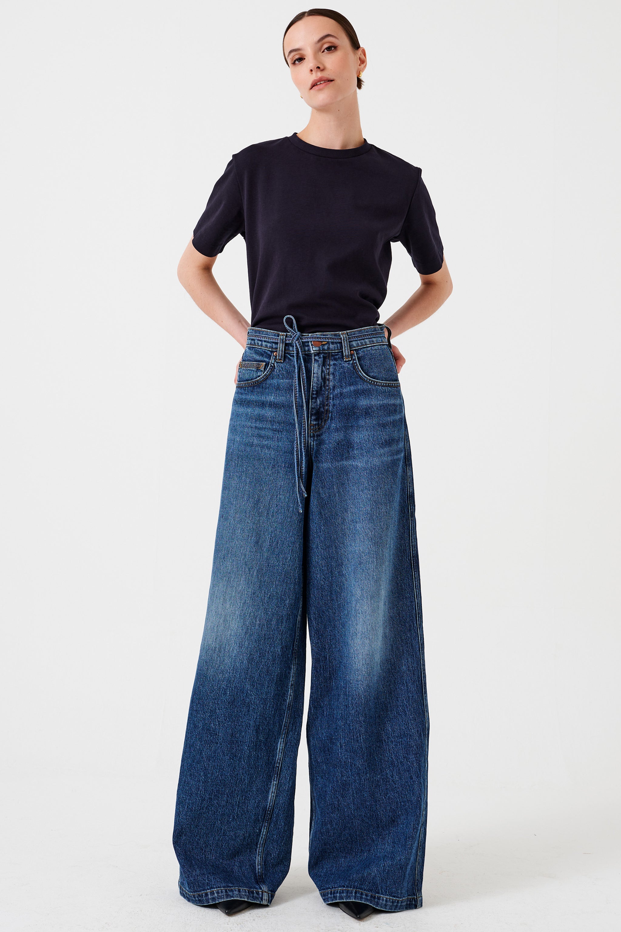 Woman wearing a black t-shirt and blue wide-leg jeans with a tie waist string detail on a white background