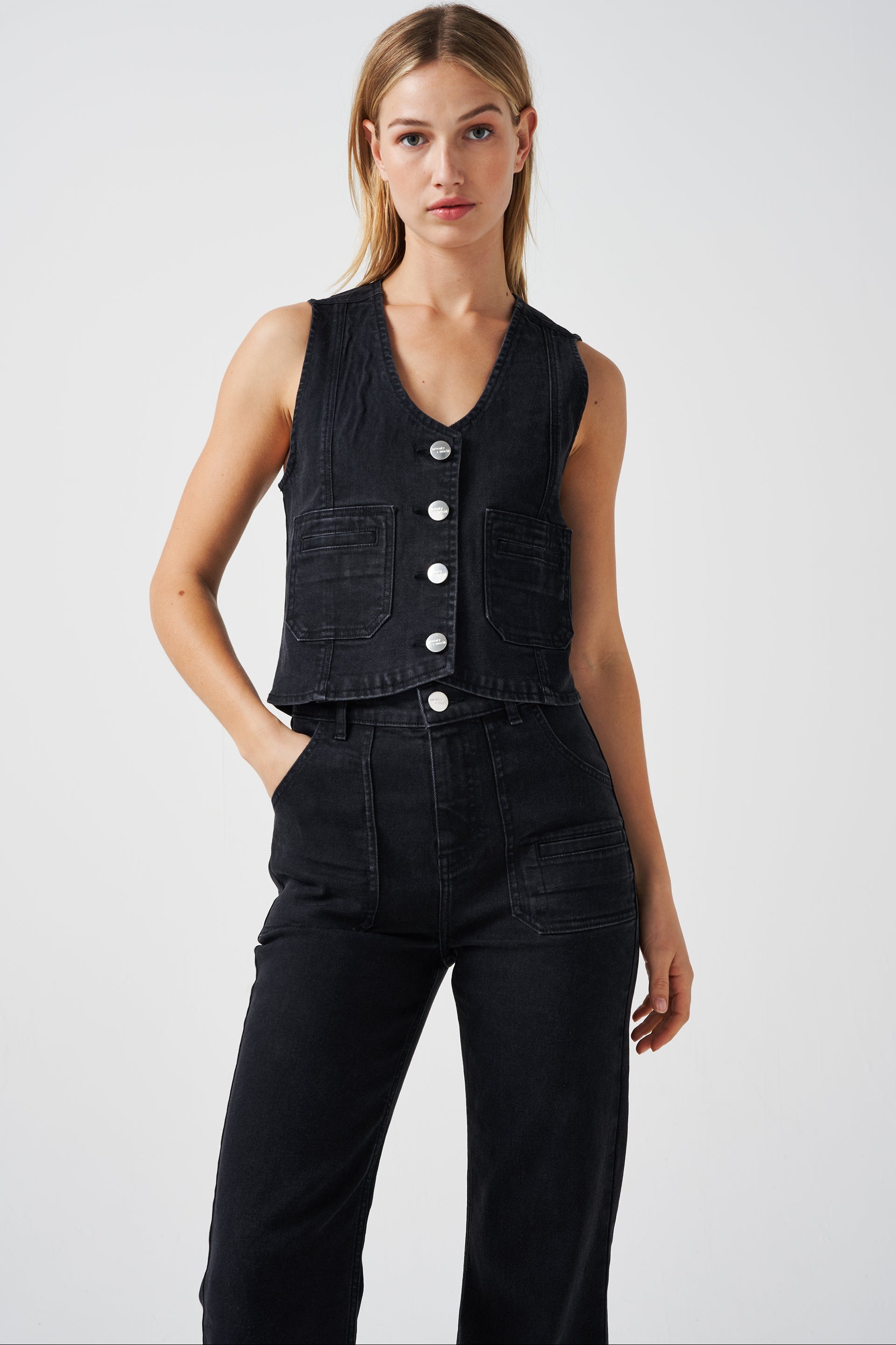woman wearing a black denim waistcoat/ vest with two large patch pockets and black jeans on a white background