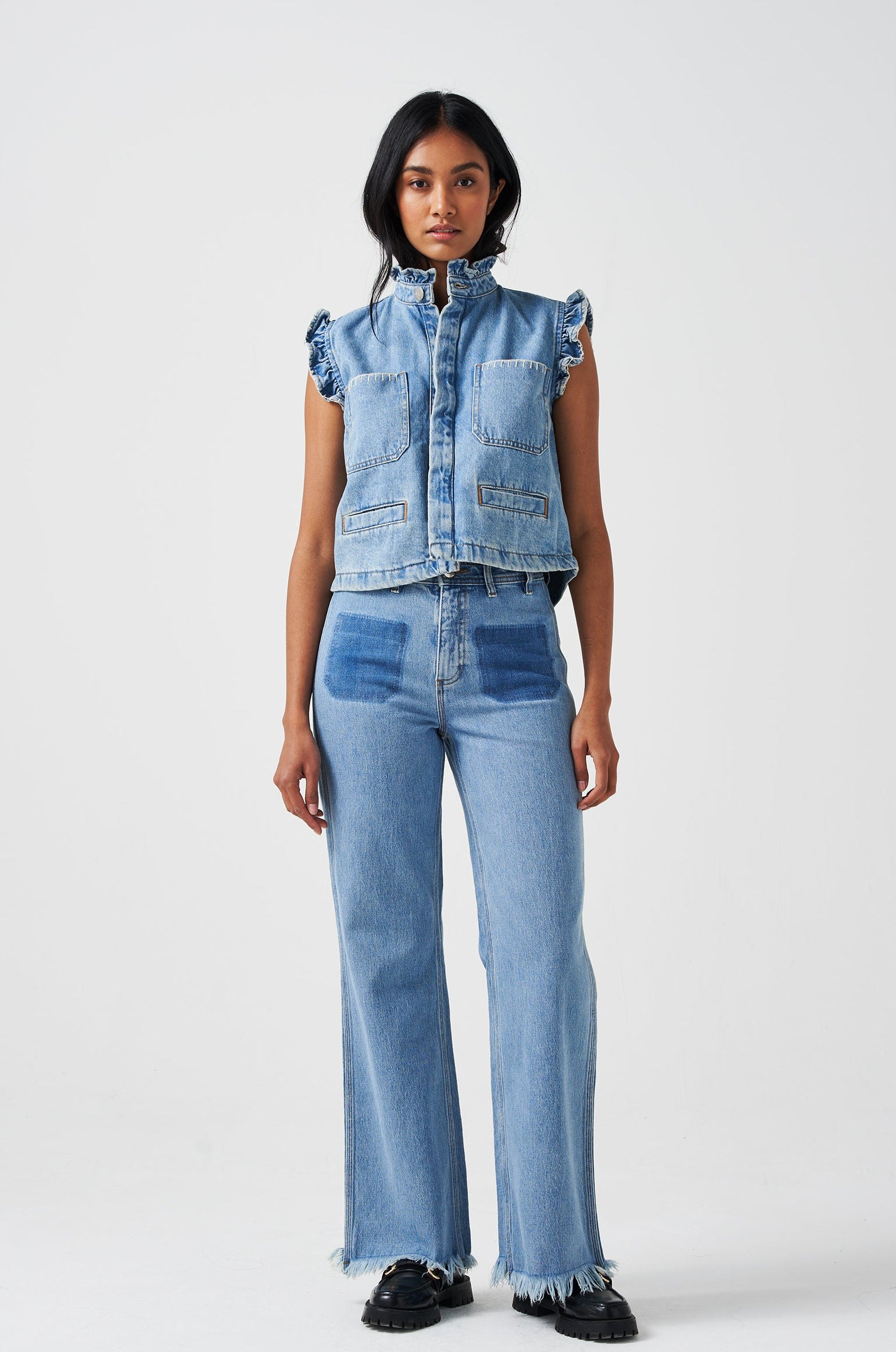 woman wearing a blue denim waistcoat/ vest with frill collar and sleeves, and two large patch pockets with white stitching, paired with blue jeans on a white background