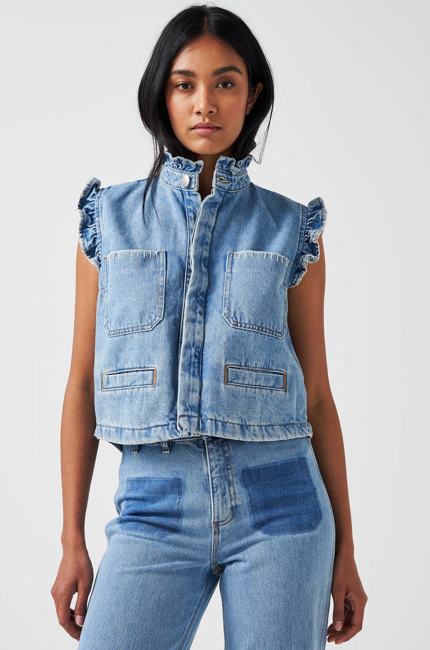 woman wearing a blue denim waistcoat/ vest with frill collar and sleeves, and two large patch pockets with white stitching, paired with blue jeans on a white background