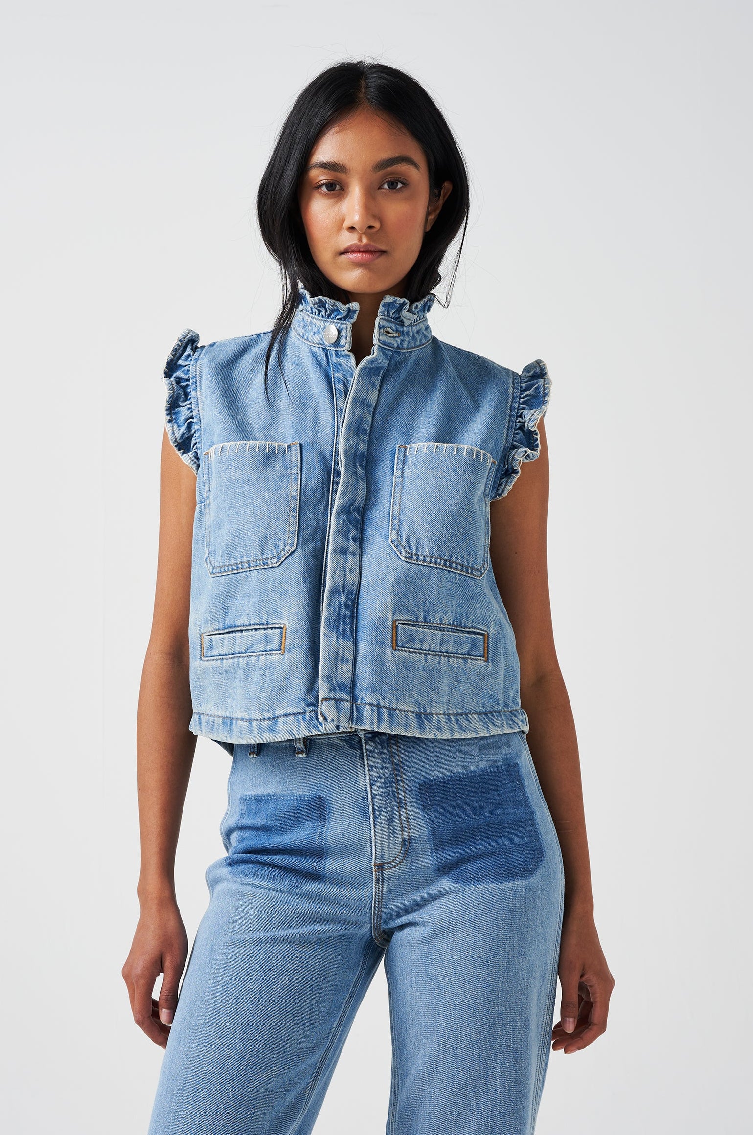 woman wearing a blue denim waistcoat/ vest with frill collar and sleeves, and two large patch pockets with white stitching, paired with blue jeans on a white background