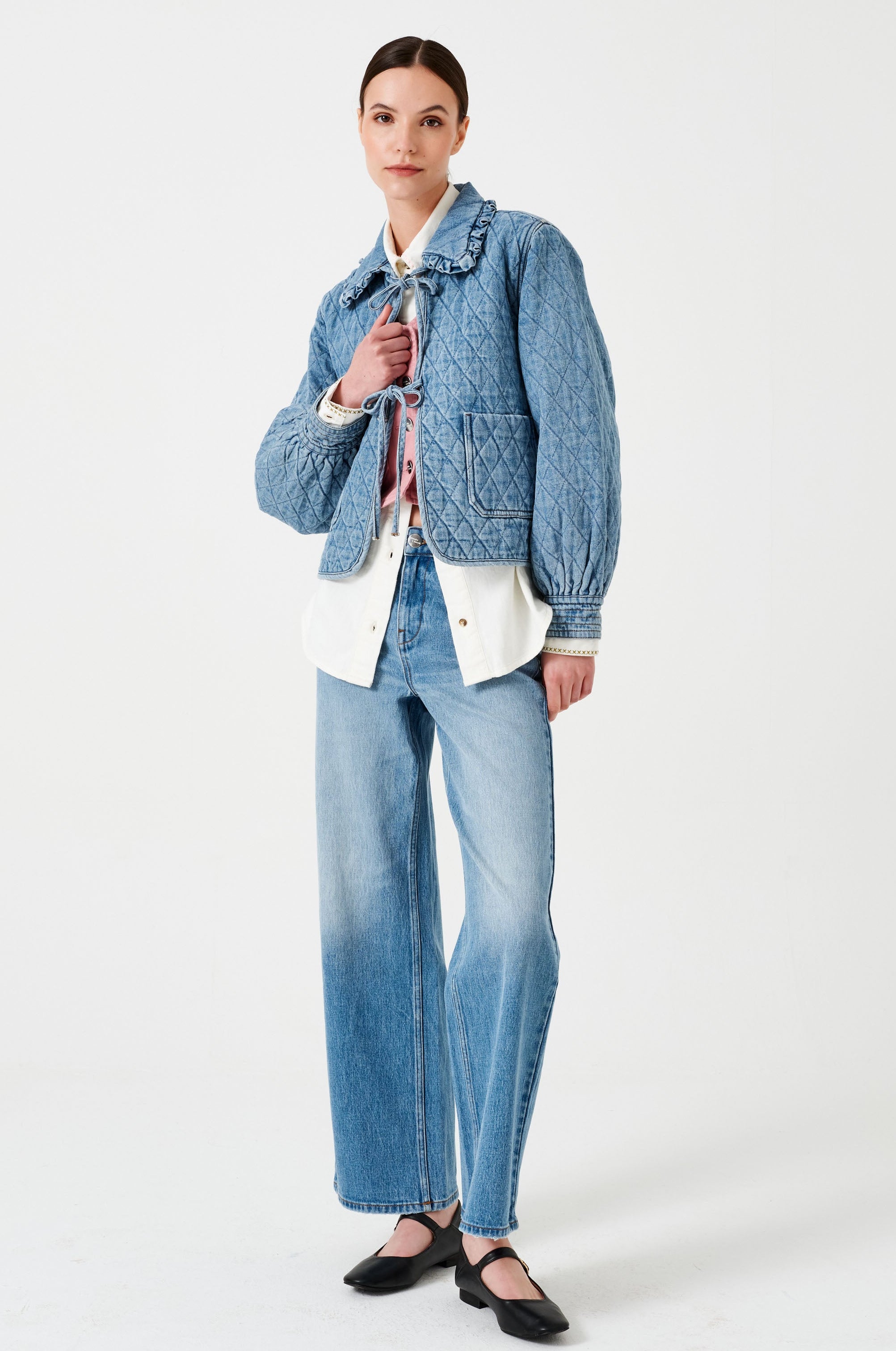 A woman wearing a light blue quilted denim jacket with a frill collar, front tie closures, and two large pockets paired with a white shirt, pink vest and blue jeans on a white background