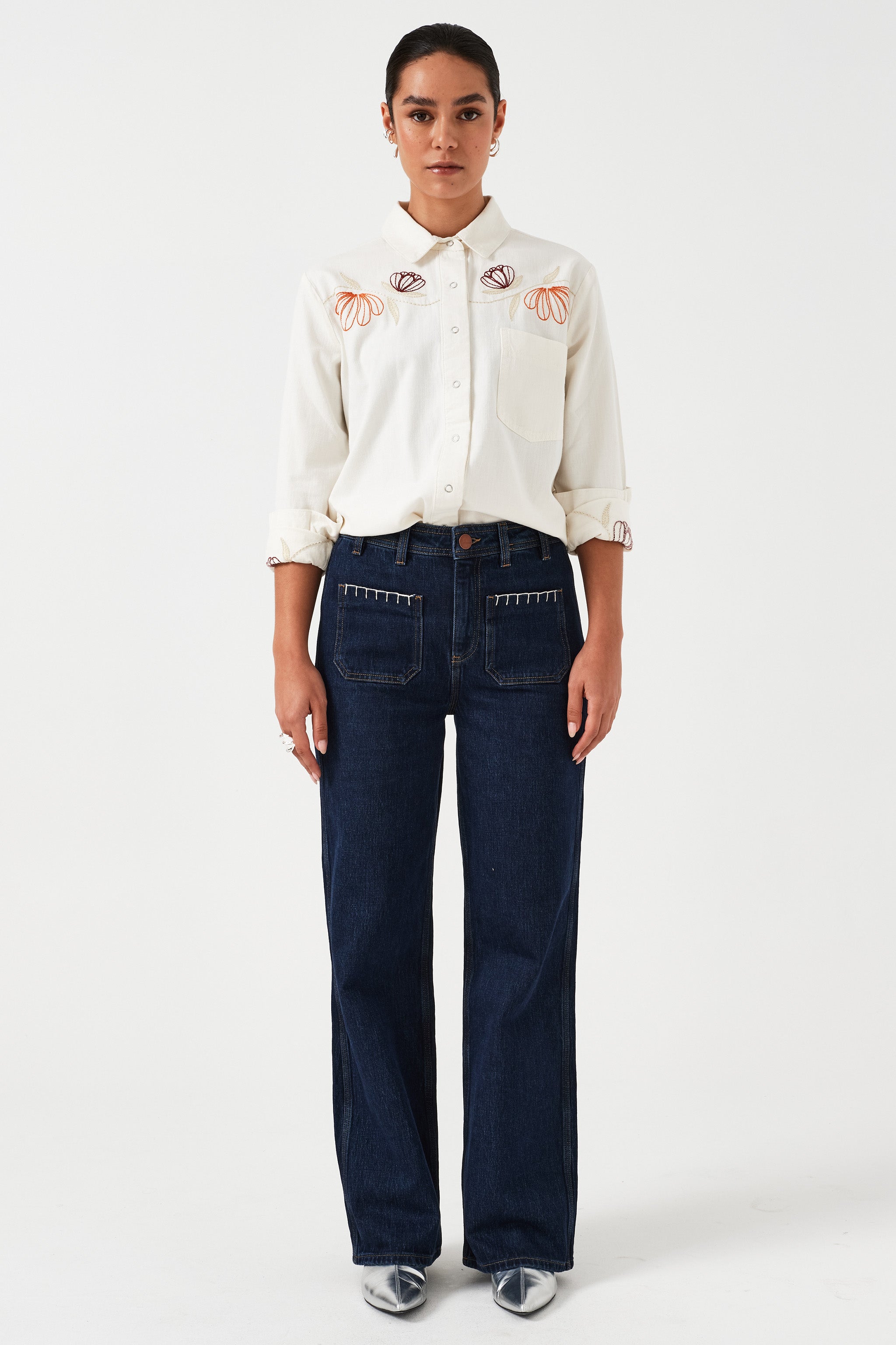 Woman standing wearing a pair of dark vintage wide-leg jeans with high-rise waist, paired with a white blouse with floral embroidery.