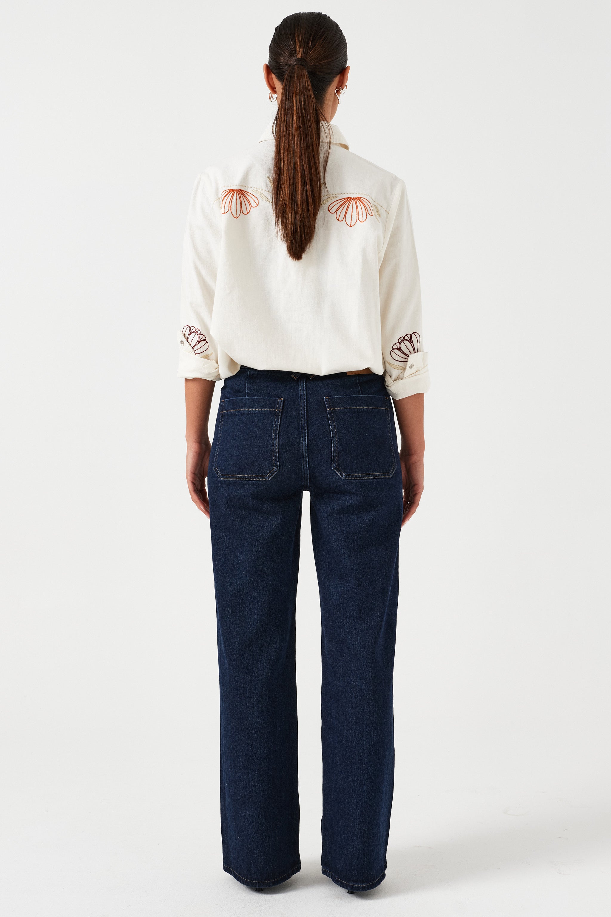 Woman standing and showing the back of a pair of dark vintage wide-leg jeans with high-rise waist, paired with a white blouse with floral embroidery.