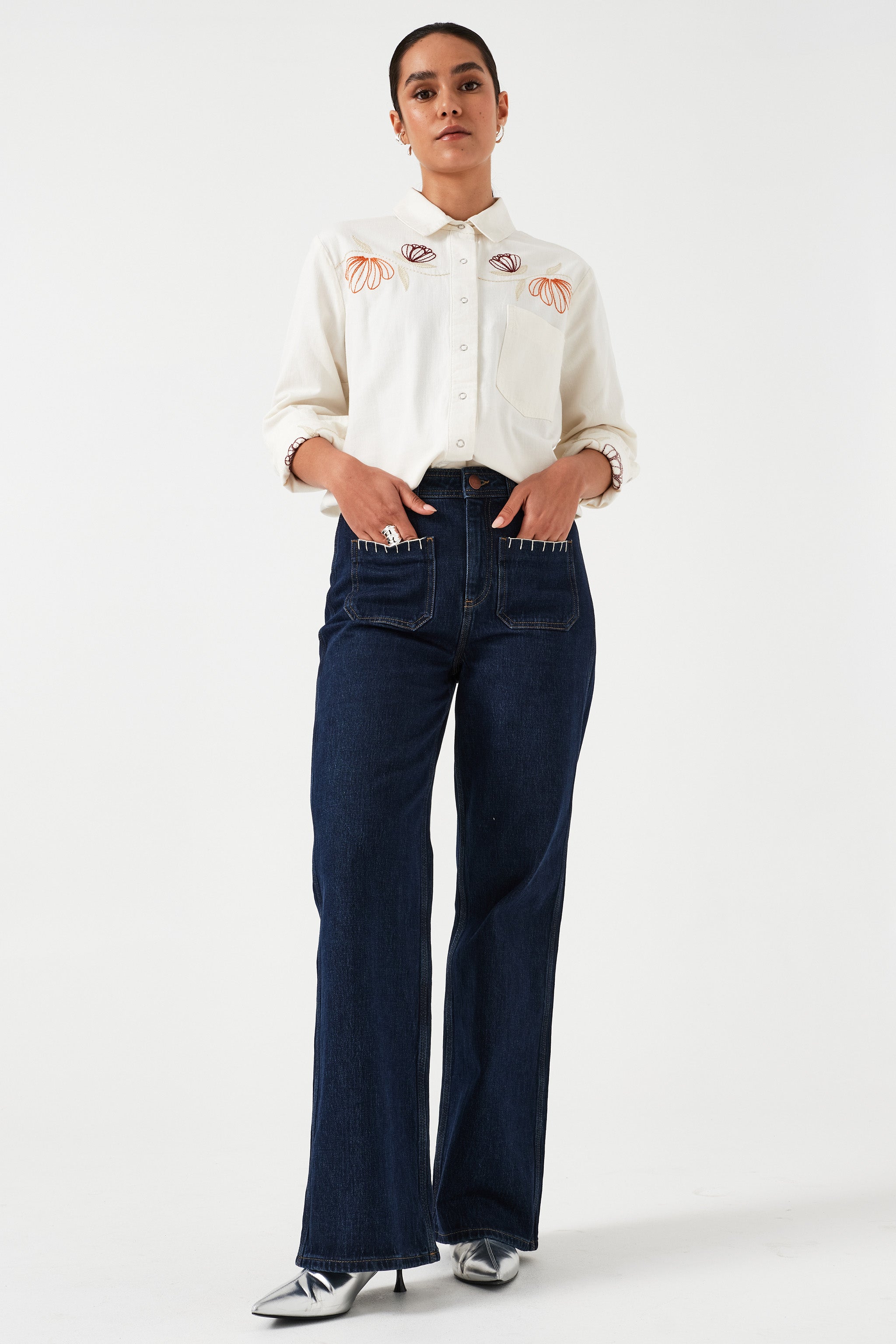Woman standing wearing a pair of dark vintage wide-leg jeans with high-rise waist, paired with a white blouse with floral embroidery.