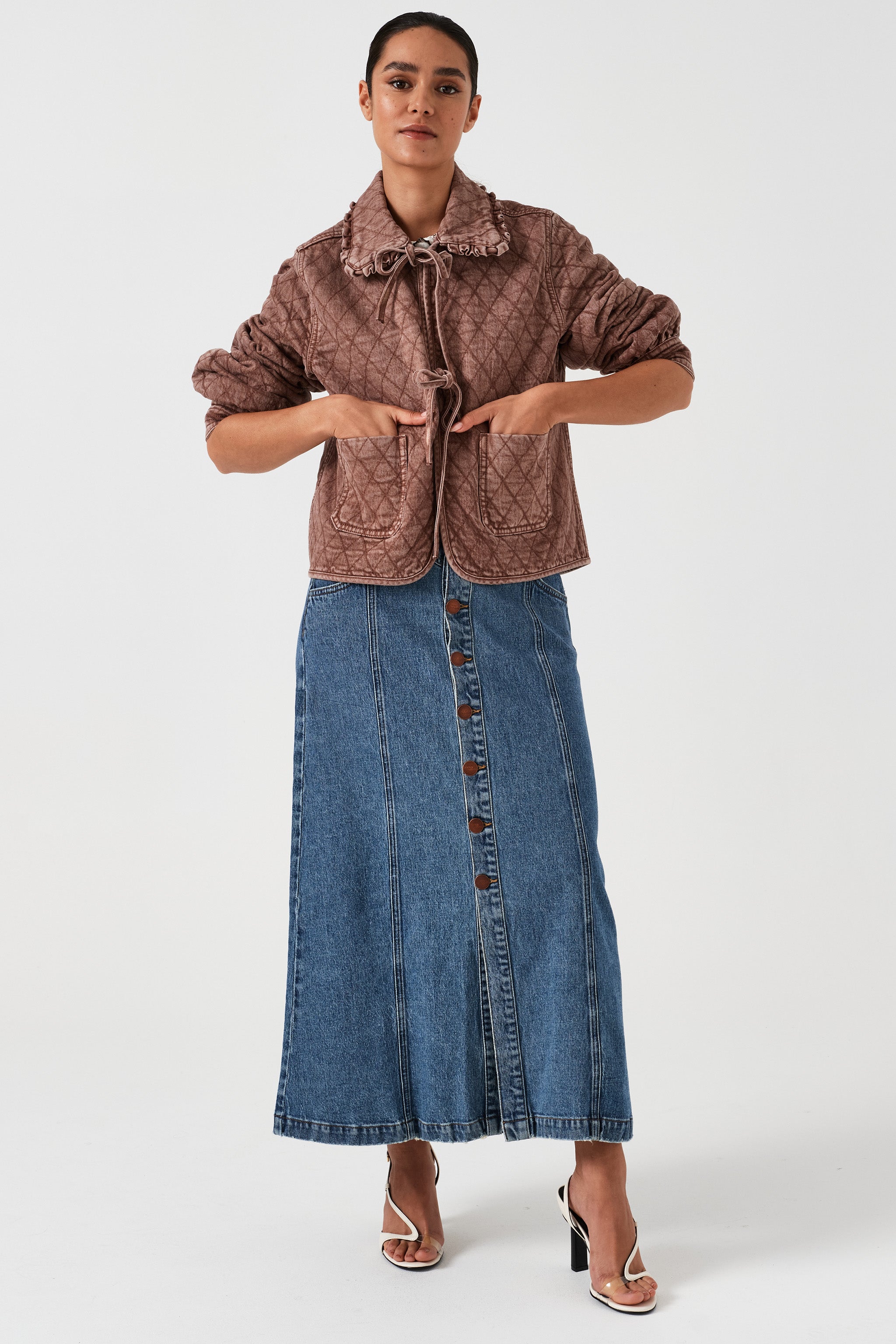 Woman wearing a brown quilted denim jacket with large pockets, a frill collar, and front tie closure, paired with a blue skirt on a white background