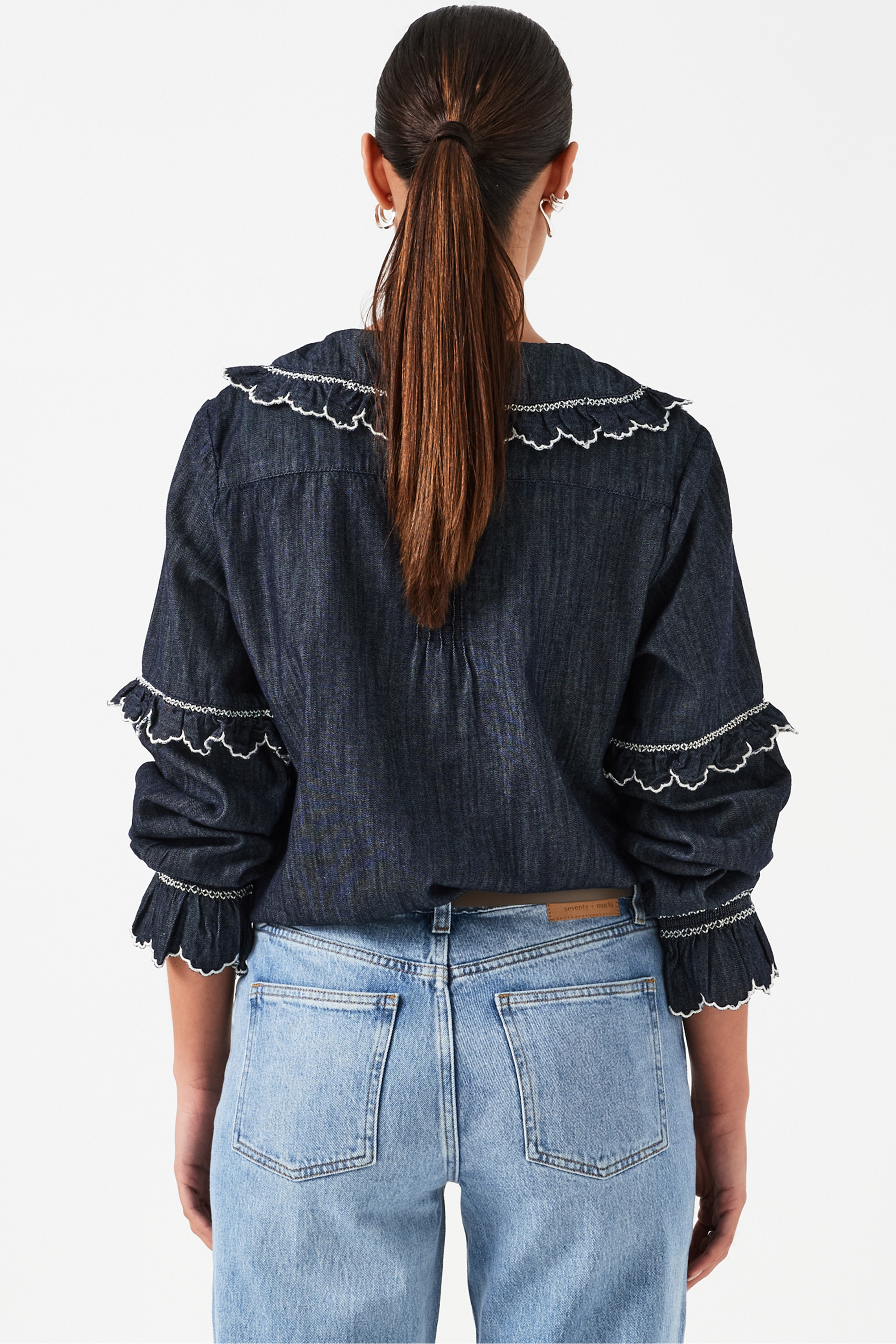 A woman wearing a navy denim blouse with a white trim frill collar and frill sleeves, paired with blue jeans on a white background. The blouse is lightweight and made of organic cotton.