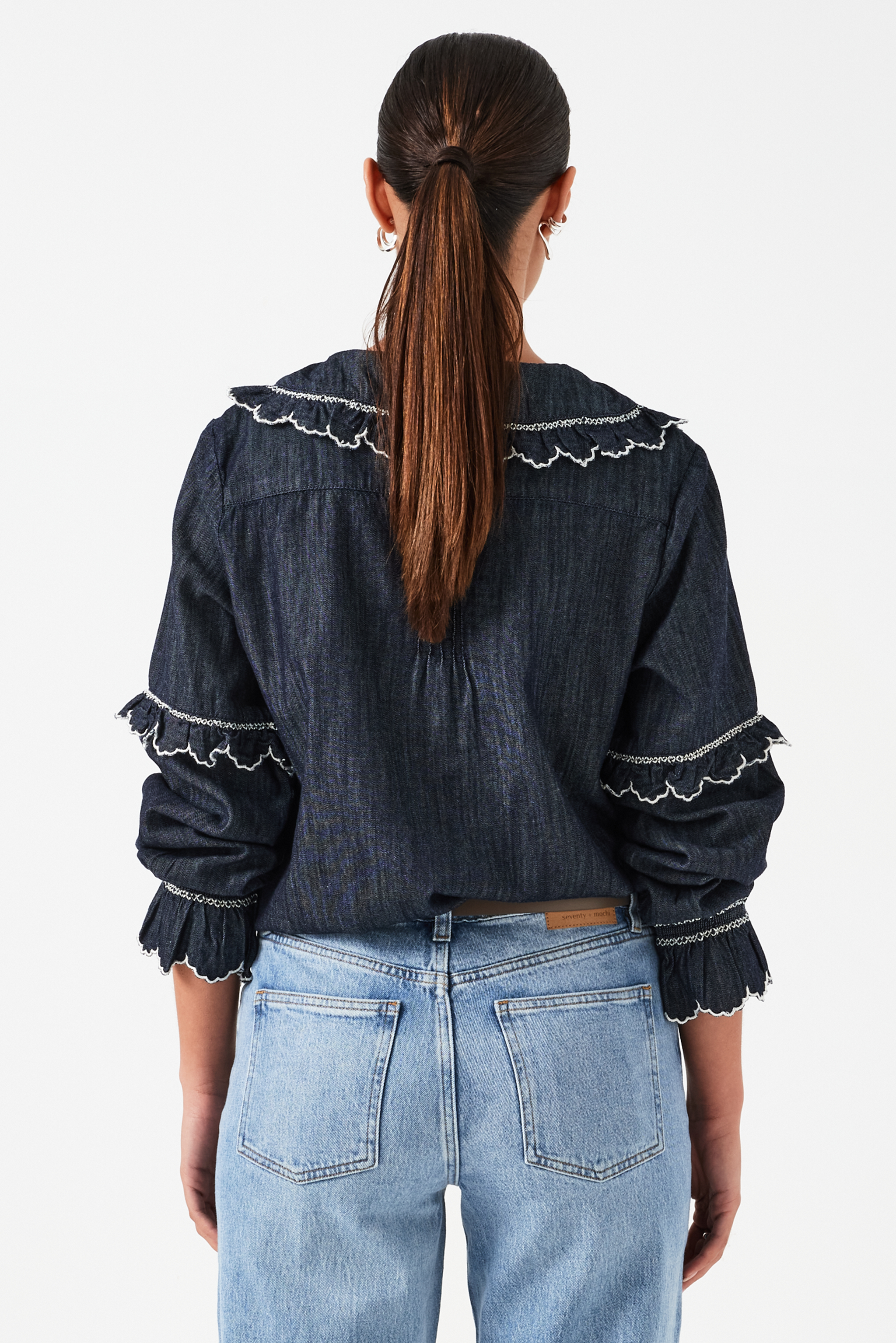 A woman wearing a navy denim blouse with a white trim frill collar and frill sleeves, paired with blue jeans on a white background. The blouse is lightweight and made of organic cotton.