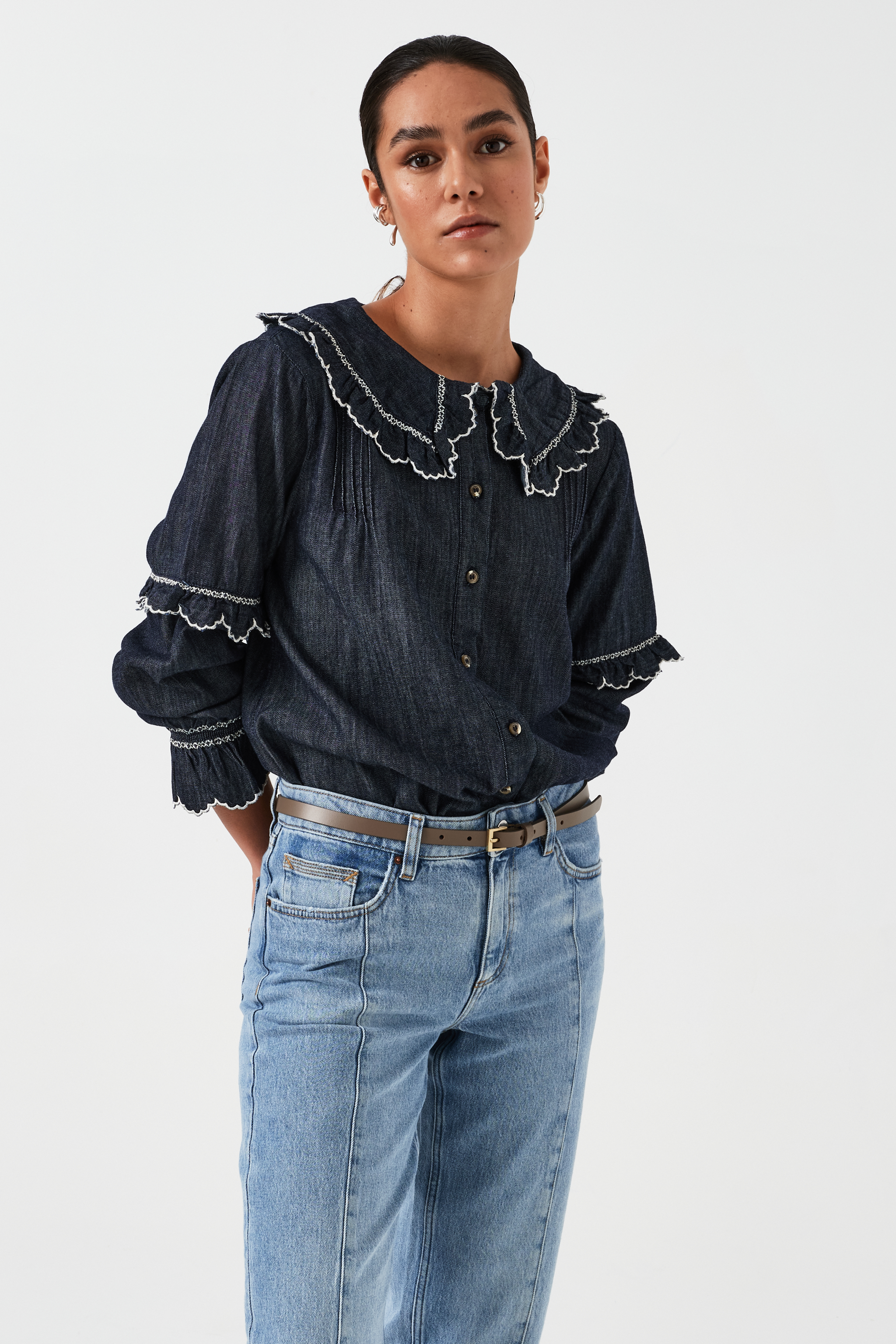 A woman wearing a navy denim blouse with a white trim frill collar and frill sleeves, paired with blue jeans on a white background. The blouse is lightweight and made of organic cotton.