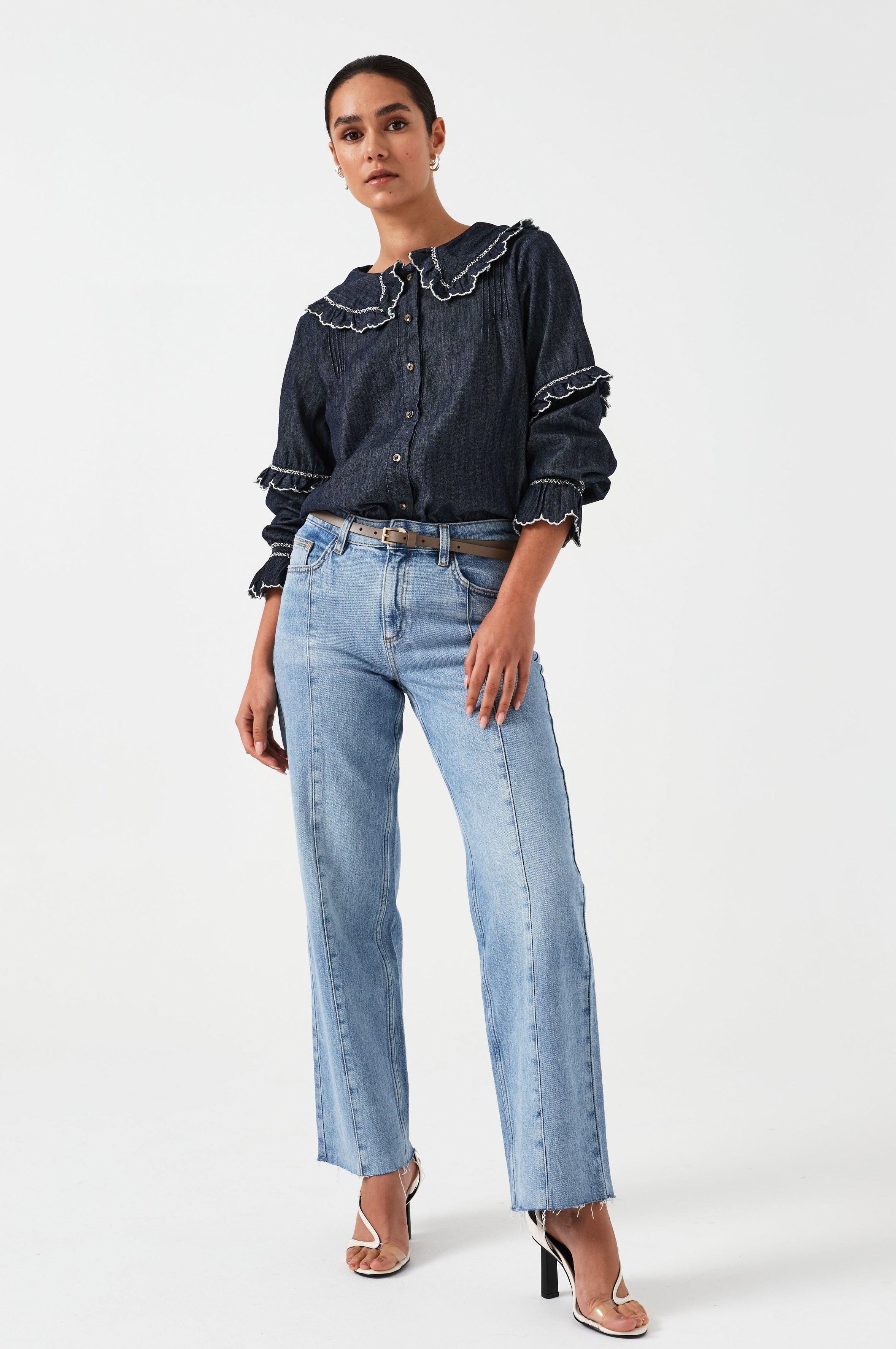A woman wearing a navy denim blouse with a white trim frill collar and frill sleeves, paired with blue jeans on a white background. The blouse is lightweight and made of organic cotton.