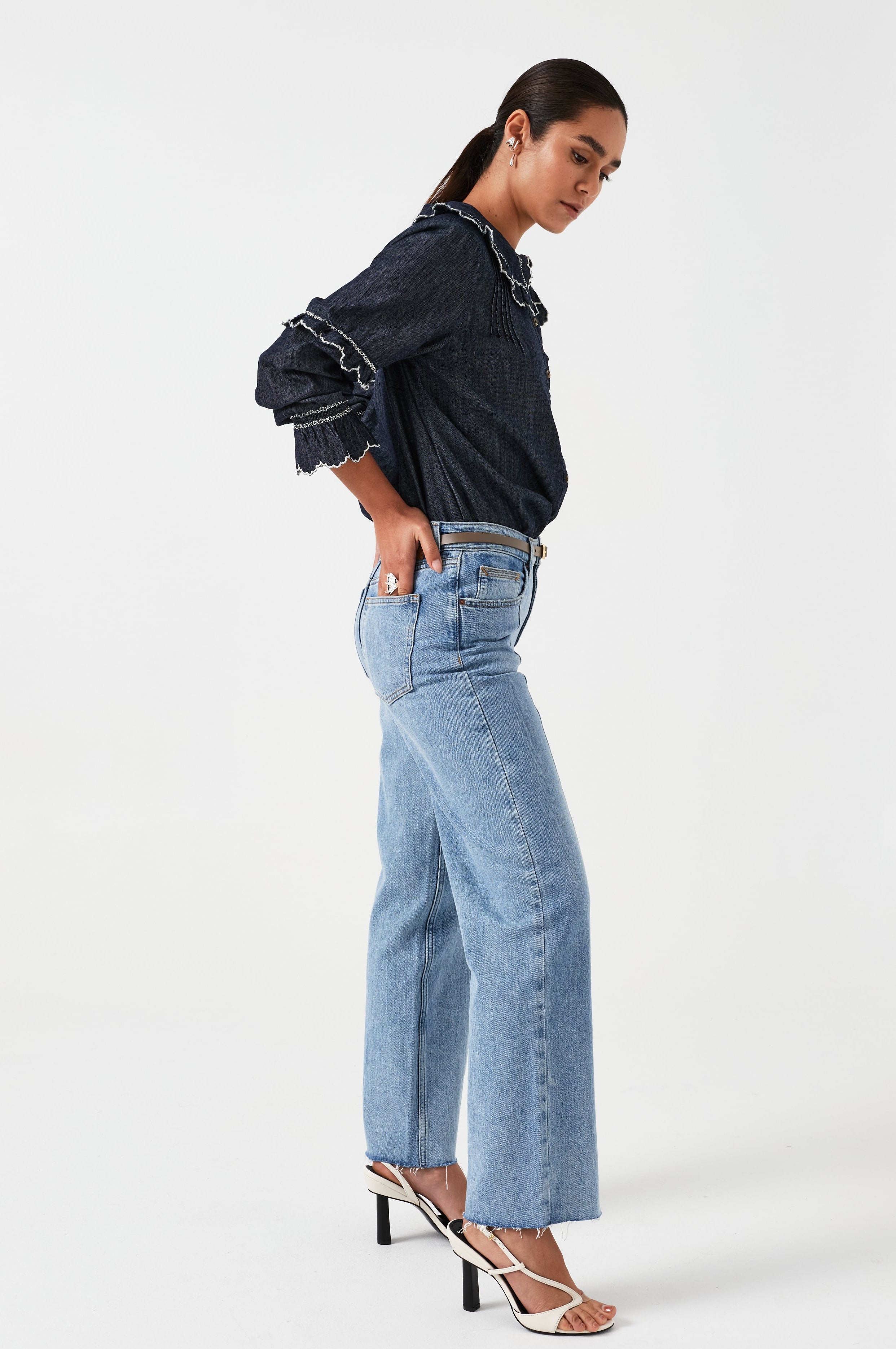 A woman wearing a navy denim blouse with a white trim frill collar and frill sleeves, paired with blue jeans on a white background. The blouse is lightweight and made of organic cotton.