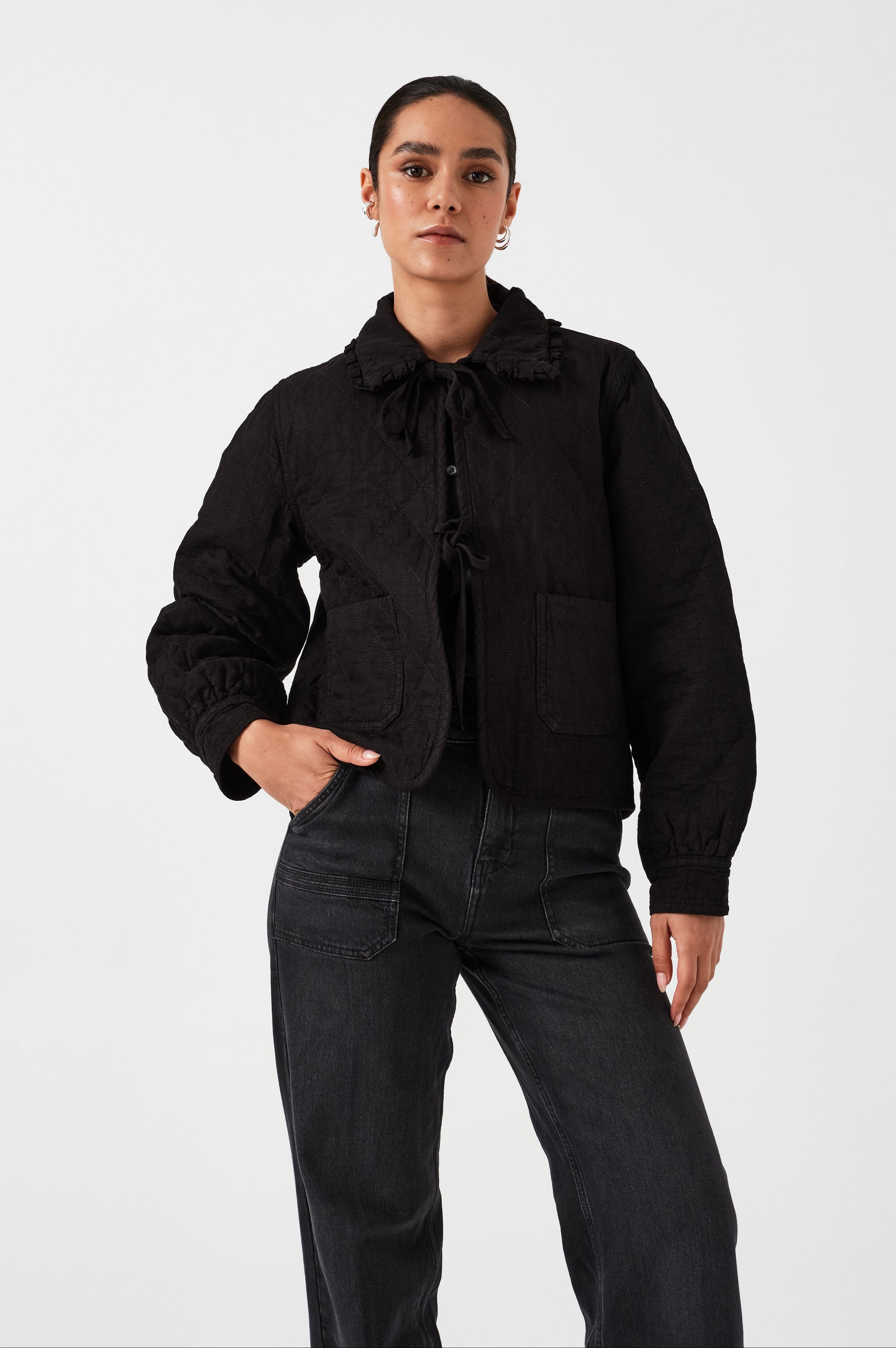 Woman wearing a black quilted denim jacket with a ruffled collar, front tie closures, and two large pockets on a white background