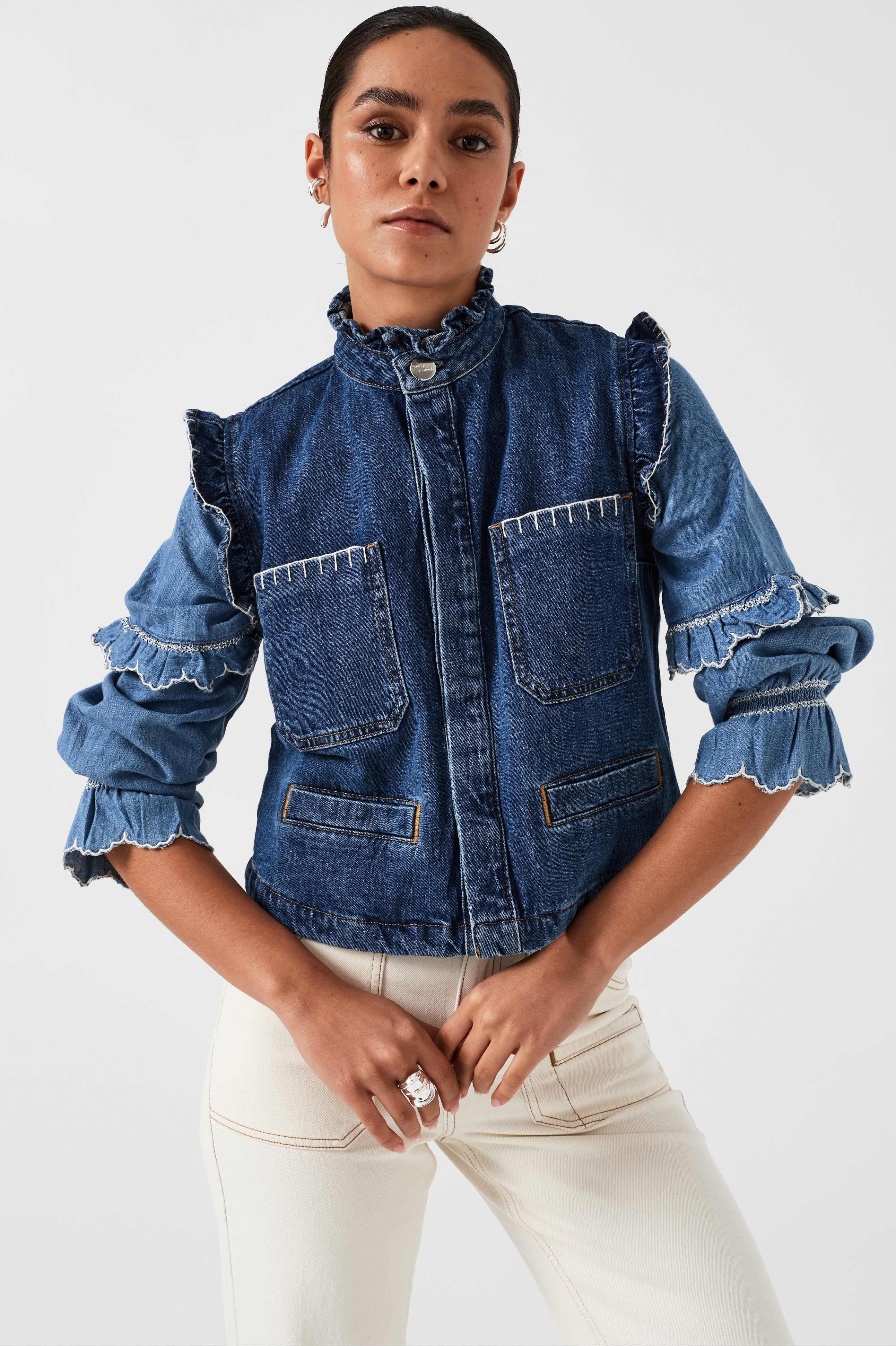 woman wearing a blue denim vest/ waistcoat with our signature white stitch detailing, two large patch pockets, and a frill collar with a blue shirt and white jeans against a white background
