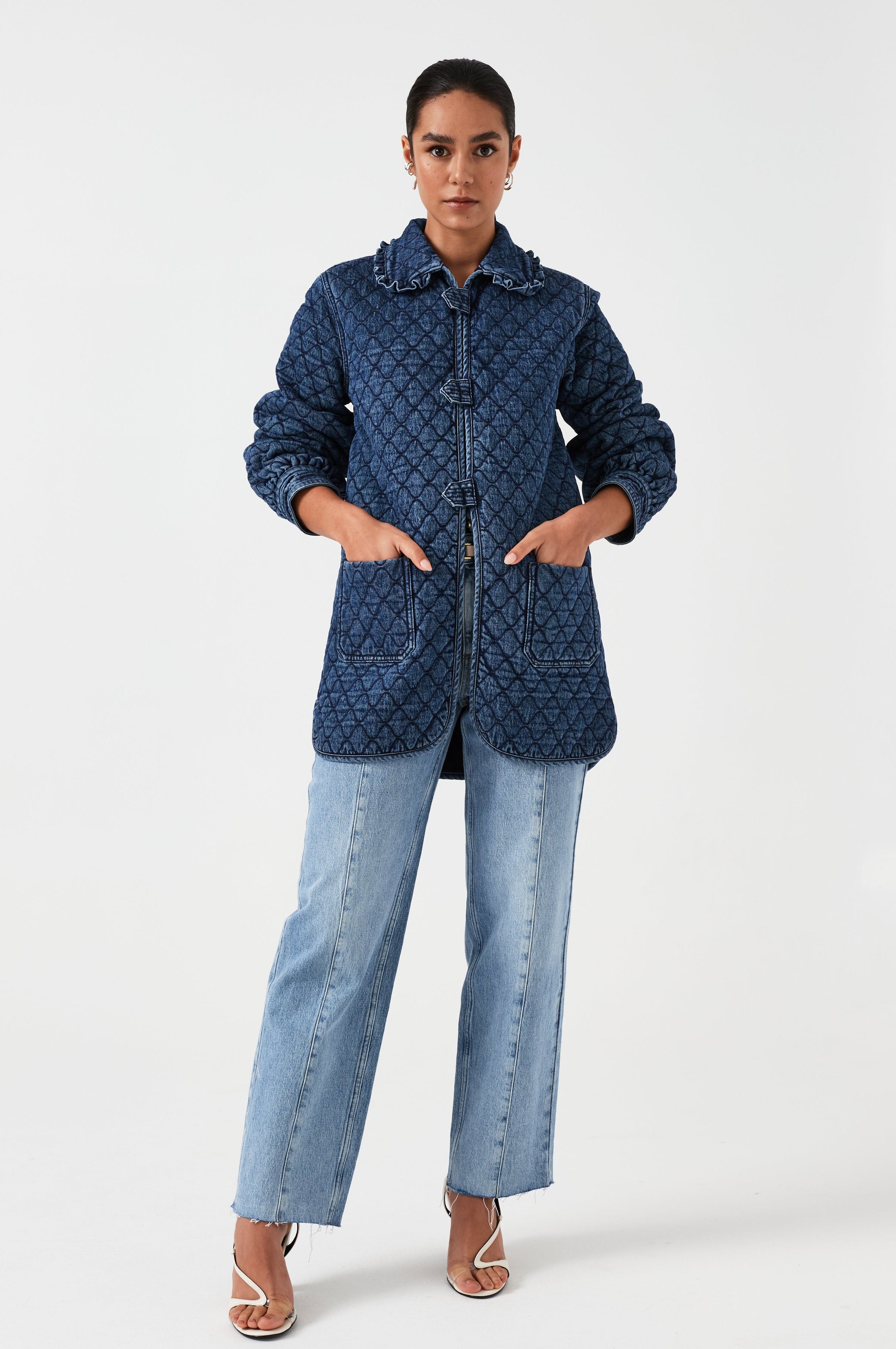 woman wearing a blue quilted jacket with a frill collar, tab closures, and two large pockets on a white background