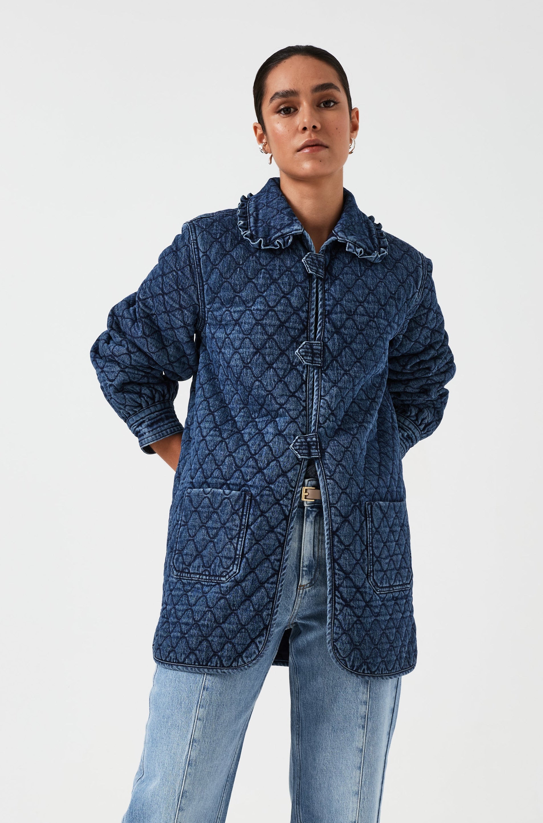 woman wearing a blue quilted jacket with a frill collar, tab closures, and two large pockets on a white background