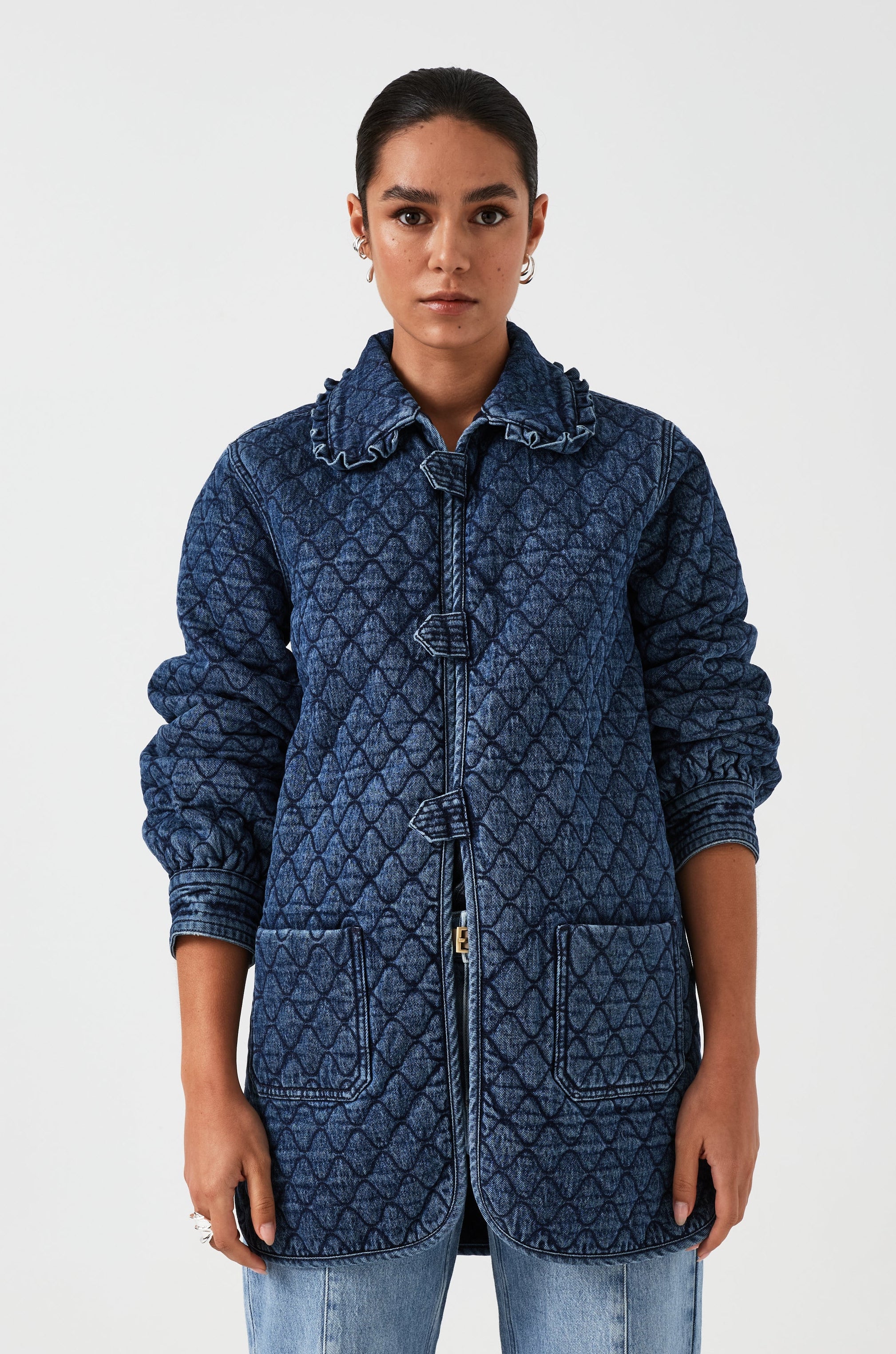 woman wearing a blue quilted jacket with a frill collar, tab closures, and two large pockets on a white background