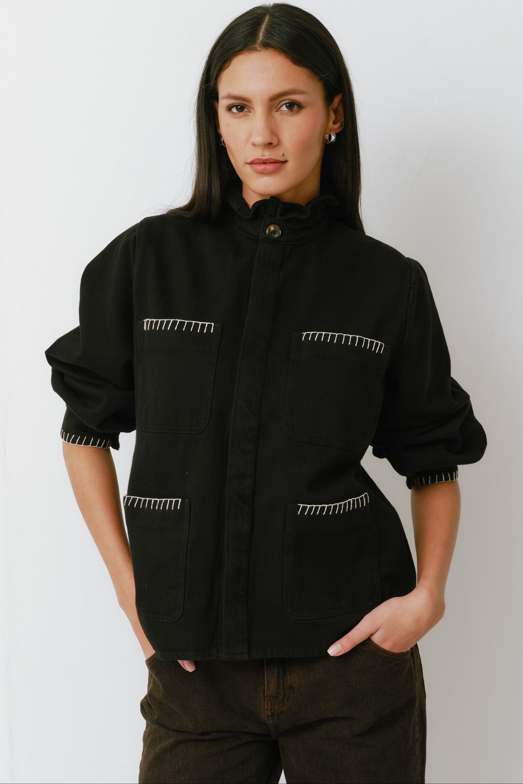 A woman wearing a black denim jacket with a frill collar and our signature white blanket stitching on four large patch pockets, paired with brown jeans against a white backdrop