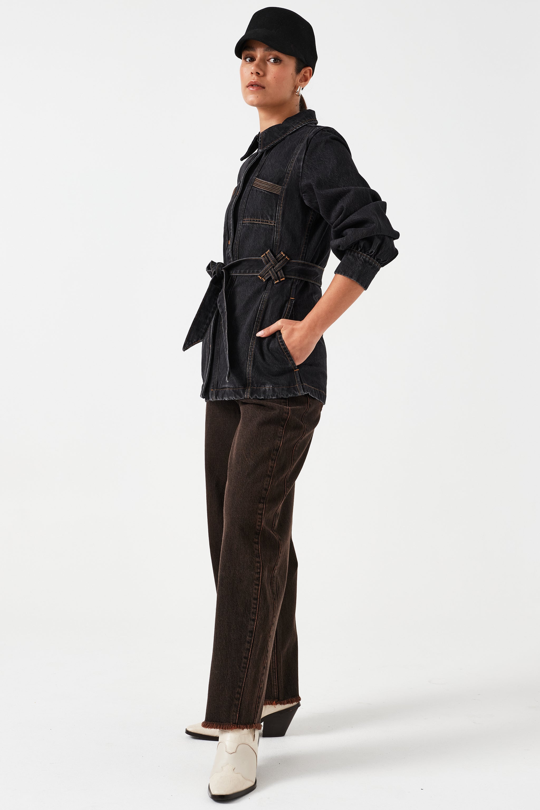 woman standing against a white background wearing high-rise dark brown cacao-colored carpenter barrel leg jeans with a frayed hem and dark wash denim jacket with a tie waist and a black hat