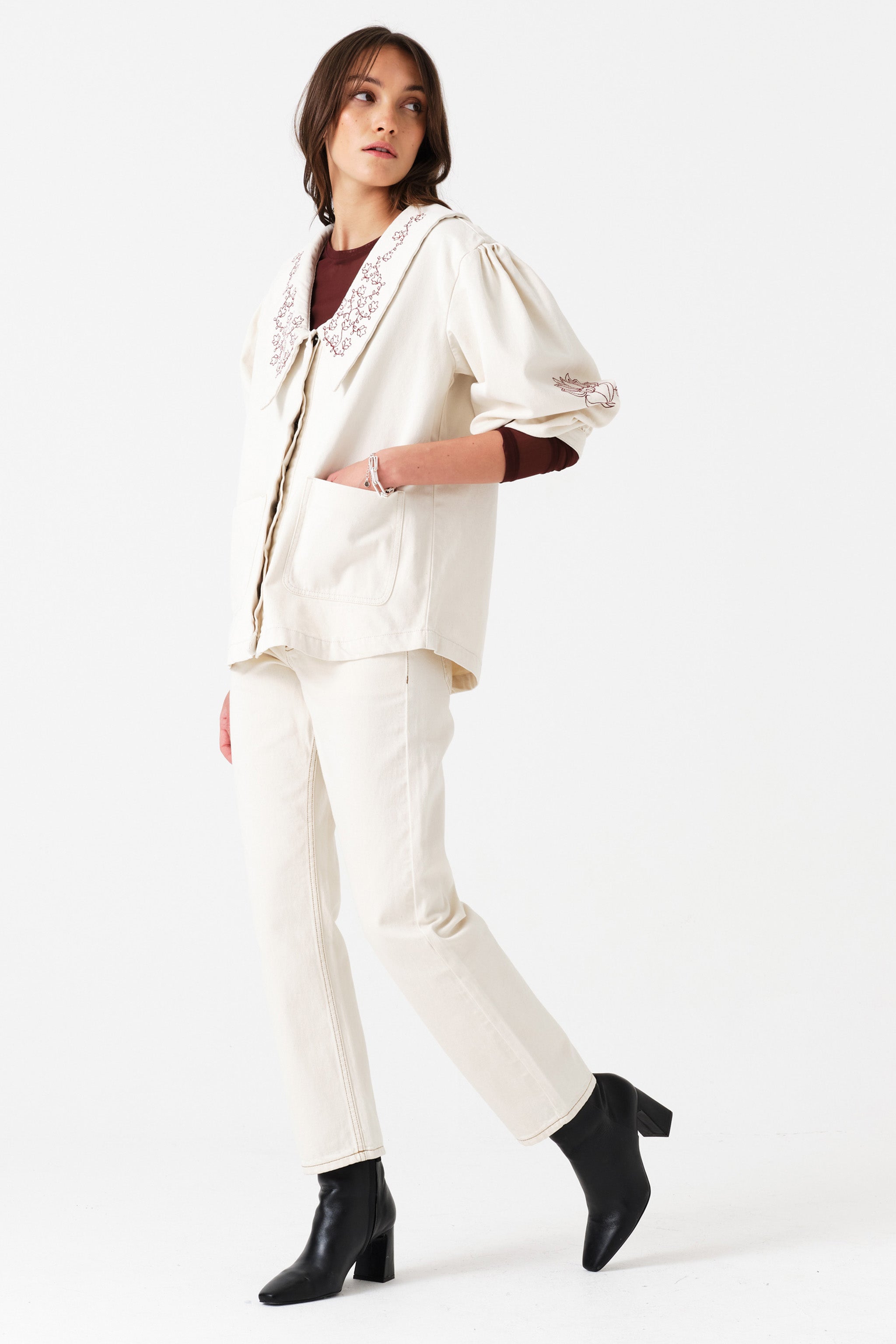Woman wearing off white straight leg jeans and an off white denim jacket with embroidered details on a white background