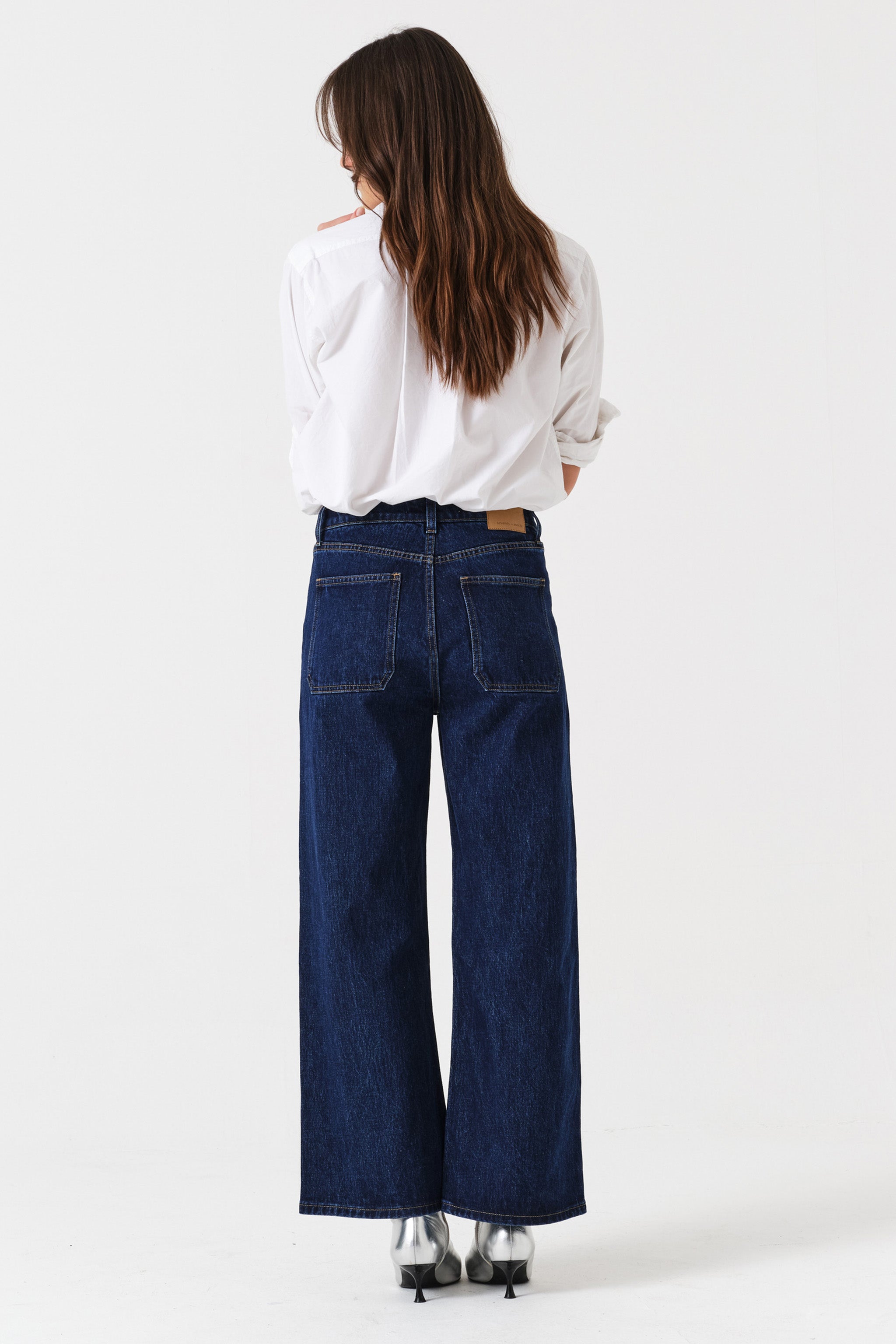 Woman wearing dark wash blue cropped wide-leg jeans and a white shirt on a white background, photographed from the back
